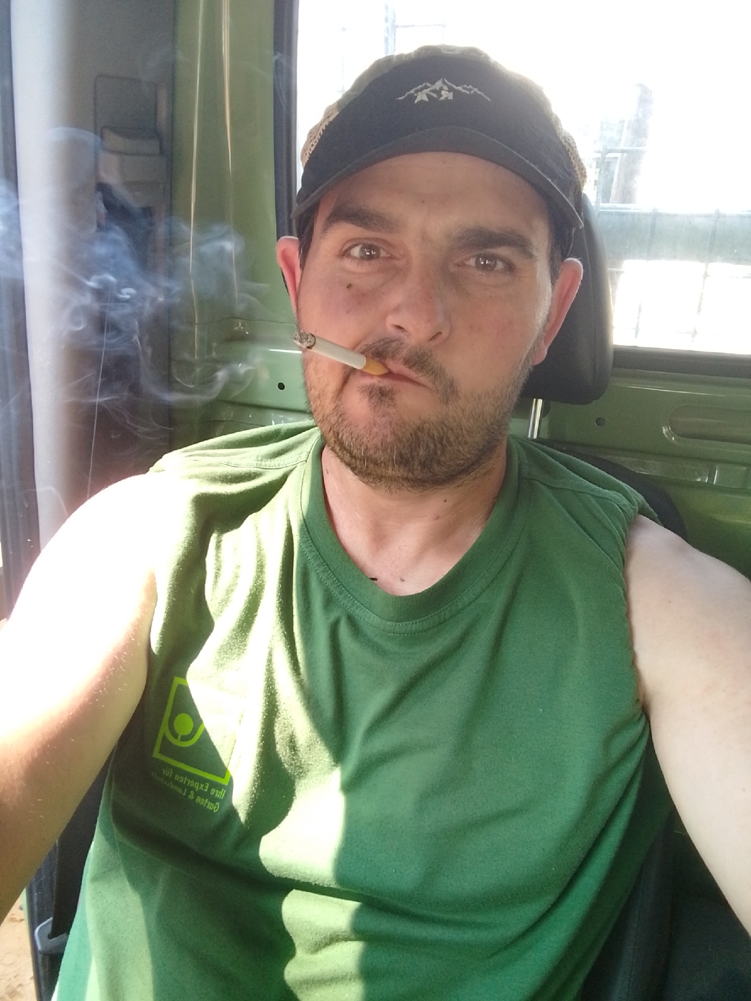 A man with a beard and a black baseball cap is sitting in what appears to be a vehicle. He is wearing a green t-shirt and smoking a cigarette, with smoke visible in the air. The man is looking directly at the camera. The background shows the interior of a vehicle with a green surface and a window with bright sunlight streaming through. There is a logo on the t-shirt that is difficult to read but appears to be a square with a circle inside and some text below it. The overall impression is a candid, close-up shot of the man. No specific location in Hamburg, Germany can be identified from the image.