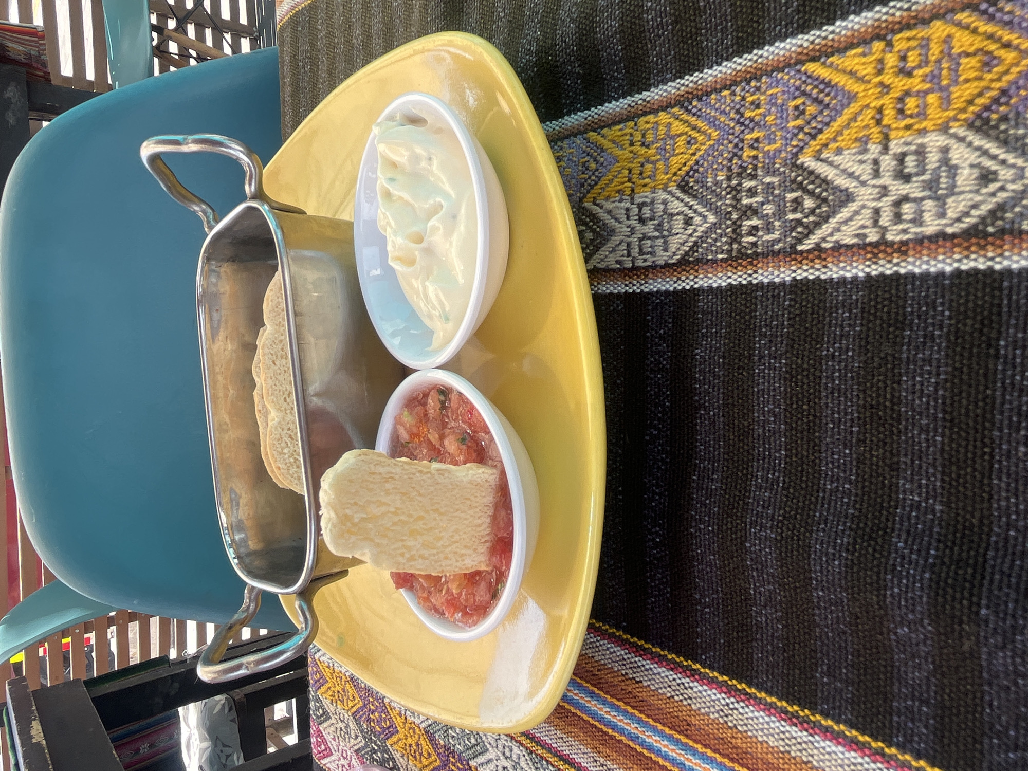 Here is a forensic description of the image:
A yellow tray holds a metal container filled with sliced bread and two small white bowls. One bowl contains a white, creamy substance, possibly mayonnaise or aioli. The other bowl holds a red mixture, likely salsa or diced tomatoes. A piece of bread is partially submerged in the red mixture. The tray rests on a tablecloth with black and yellow woven patterns. Part of a blue chair is visible on the left side of the image. The location is unknown, but appears to be outdoors.