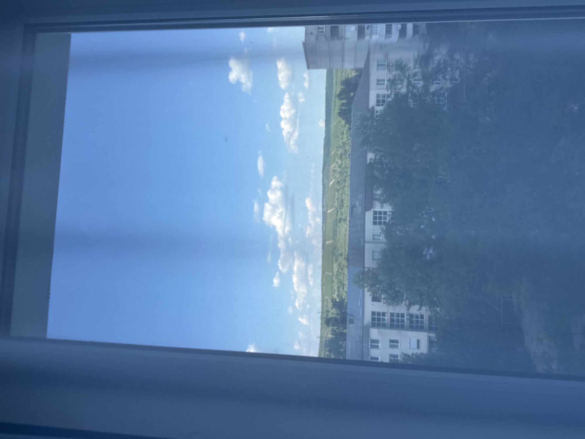 The image shows a view through a window, likely from an apartment building in Strășeni, Moldova. The orientation of the image suggests it was taken vertically. The scene depicts a bright, clear day with a vibrant blue sky dotted with fluffy white clouds. In the middle ground, there is a lush green landscape, possibly fields or a park, stretching towards the horizon. On the right side of the image, there are multi-story buildings, characteristic of an urban or suburban environment. Trees with green foliage are visible, partially obscuring some of the buildings. The buildings appear to be residential or office blocks, with visible windows. The overall impression is one of a pleasant, sunny day. There is no visible text in the image, nor are there any people or specific activities depicted. The light suggests it is daytime, likely mid-morning or afternoon, given the brightness and the angle of the sun. The view out of the window implies the photographer is indoors, looking out.