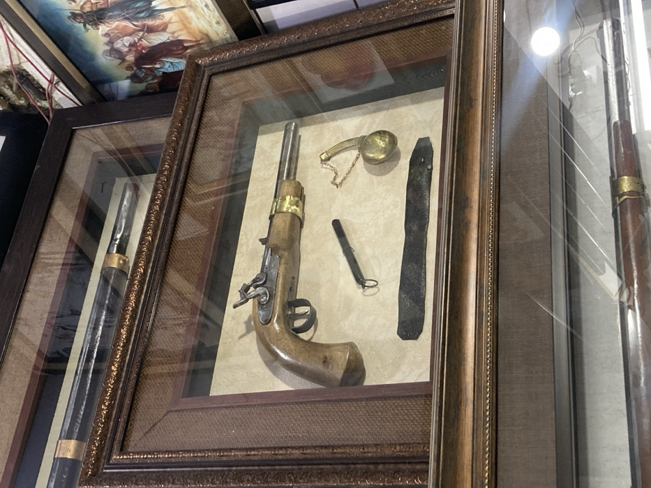 The image displays an antique weapons collection in Quetta City Tehsil, Pakistan. The primary subject is a wooden-framed, glass-fronted display case featuring a flintlock pistol and associated accessories. The pistol, positioned centrally, has a light-colored wooden stock, a metallic barrel with a brass band near the muzzle, and a visible flintlock mechanism. To the lower left of the pistol lies a dark, two-pronged tool, possibly a pick, with two small rings. To the lower right is a brass-toned powder flask with an attached chain. Below these items, a dark, textured strap or belt section is arranged horizontally. The background of this display case is a light, neutral fabric.
Above this main display, a portion of another similar framed display case is visible, containing the barrel section of what appears to be a long firearm, also with a brass accent. To the right of the stacked display cases, a framed painting is partially visible, depicting multiple figures in historical attire, some mounted on horseback, rendered in warm earth tones. Reflections are present on the glass surfaces of the display cases. No individuals are visible in the scene, and no actions or interactions are depicted. The environment appears to be an indoor setting, likely a museum, collection, or retail space.