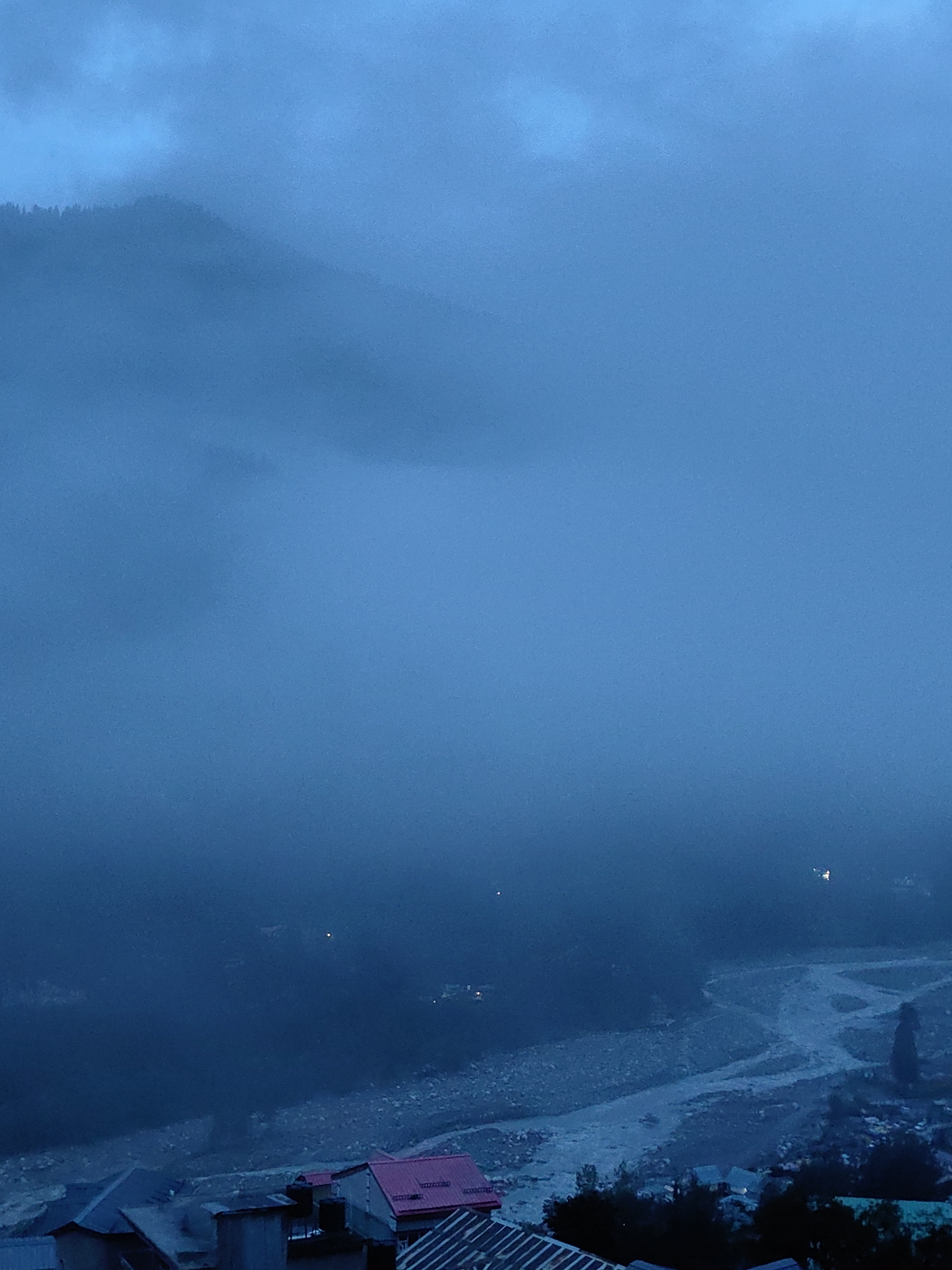 The image depicts a mountainous landscape shrouded in heavy fog, suggesting a twilight or pre-dawn setting. In the foreground, a cluster of buildings with distinct red and dark-colored roofs are visible, characteristic of traditional architecture in Vashisht, India. The dense fog obscures the visibility of the surrounding environment, including the river valley and distant mountains, which are only faintly discernible as dark shapes against the hazy sky. Scattered lights are faintly visible in the fog-laden distance, indicating human habitation. The overall atmosphere is somber and mysterious due to the low visibility and muted blue tones of the scene.