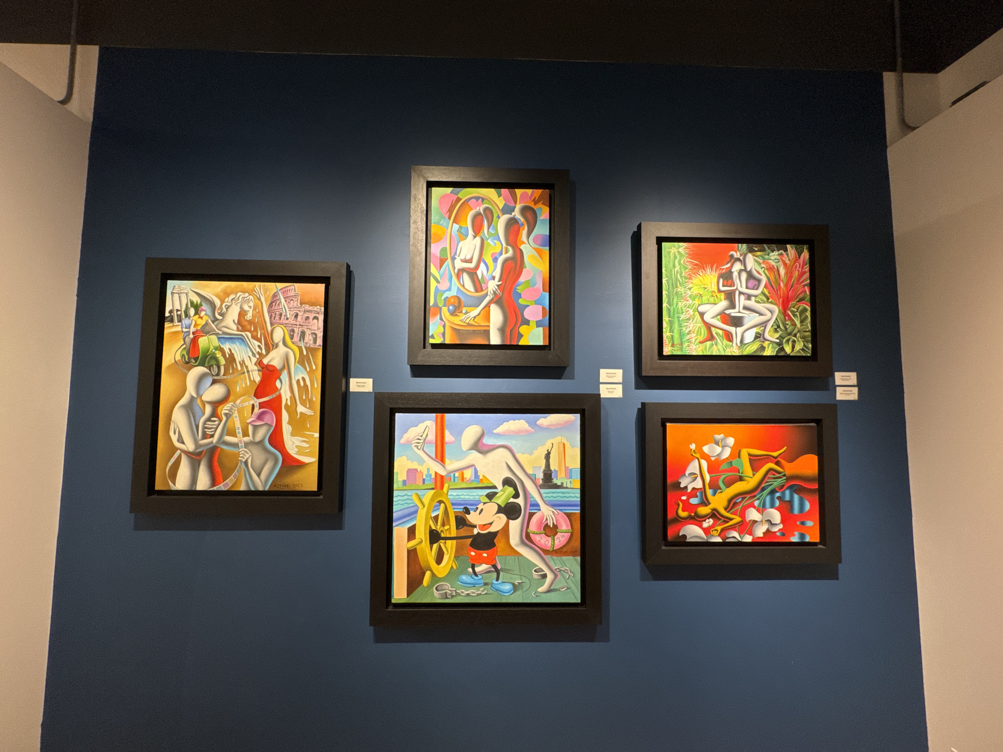The image shows five paintings displayed on a dark blue wall in what appears to be an art gallery. To the left and right of the dark blue wall are sections of white walls. The gallery is located in Schaumburg, United States of America.
The paintings are arranged in two rows. The top row consists of three paintings, and the bottom row consists of two paintings. All paintings are framed in black.
The painting on the top left depicts figures on a Vespa, seemingly in Italy, with the Colosseum in the background. The central painting on top shows two figures in front of a mirror. The painting on the top right displays a yoga pose amidst green plants. The painting on the bottom left features Mickey Mouse steering a boat with a large white figure. The painting on the bottom right depicts a figure amongst white flowers and flowing water.
Below each painting, a small white card is visible, likely containing the artist's name and the title of the work.