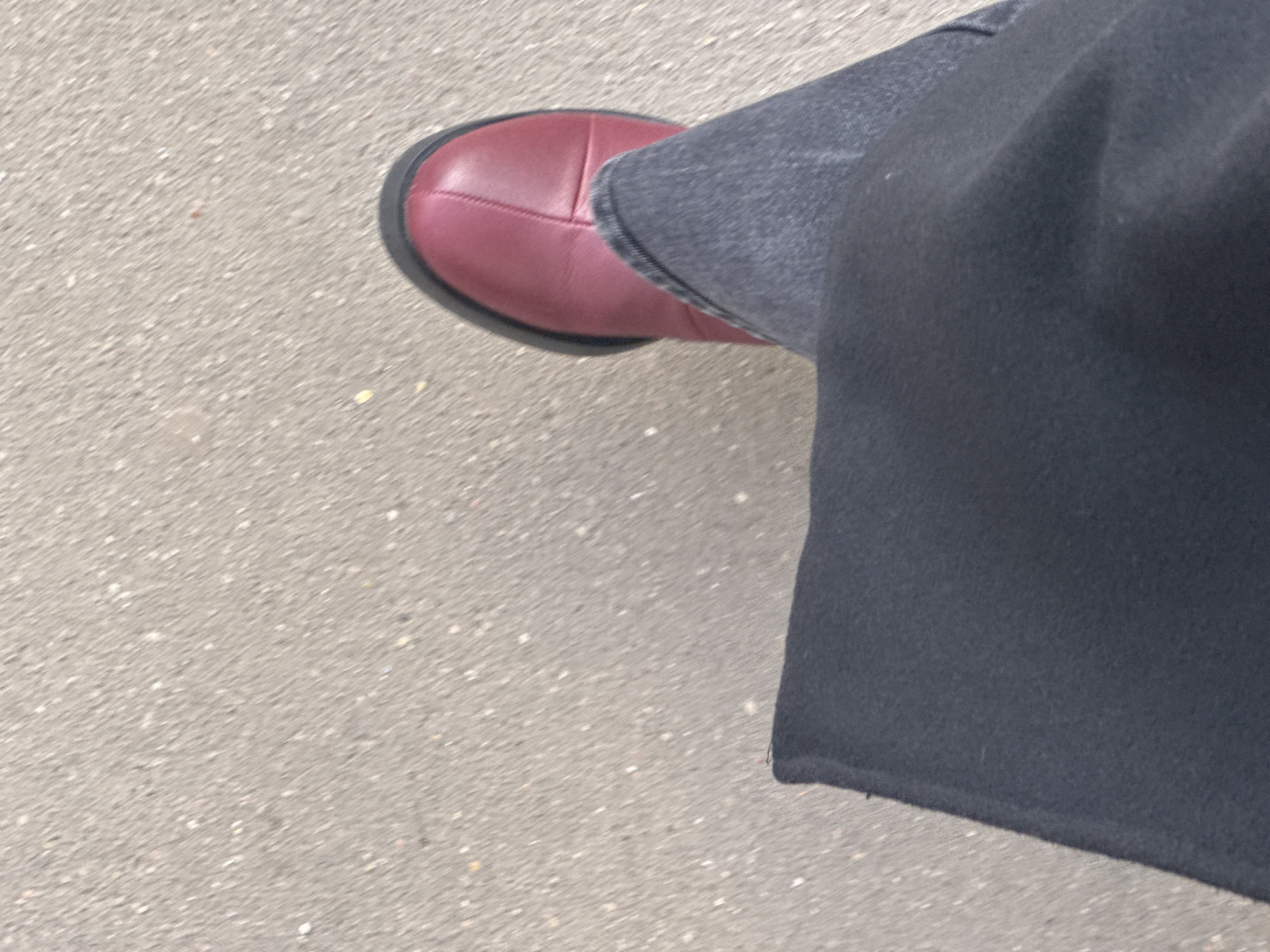 A top-down shot shows a person's left foot on a grey asphalt surface. The foot is clad in a maroon leather shoe with a black sole. The lower portion of the person's left leg is visible, covered in dark grey denim jeans and black fabric, possibly a coat. The image seems to be taken outdoors in an urban setting, but without additional visual clues, specifying the exact location within Moscow, Russia, is impossible.