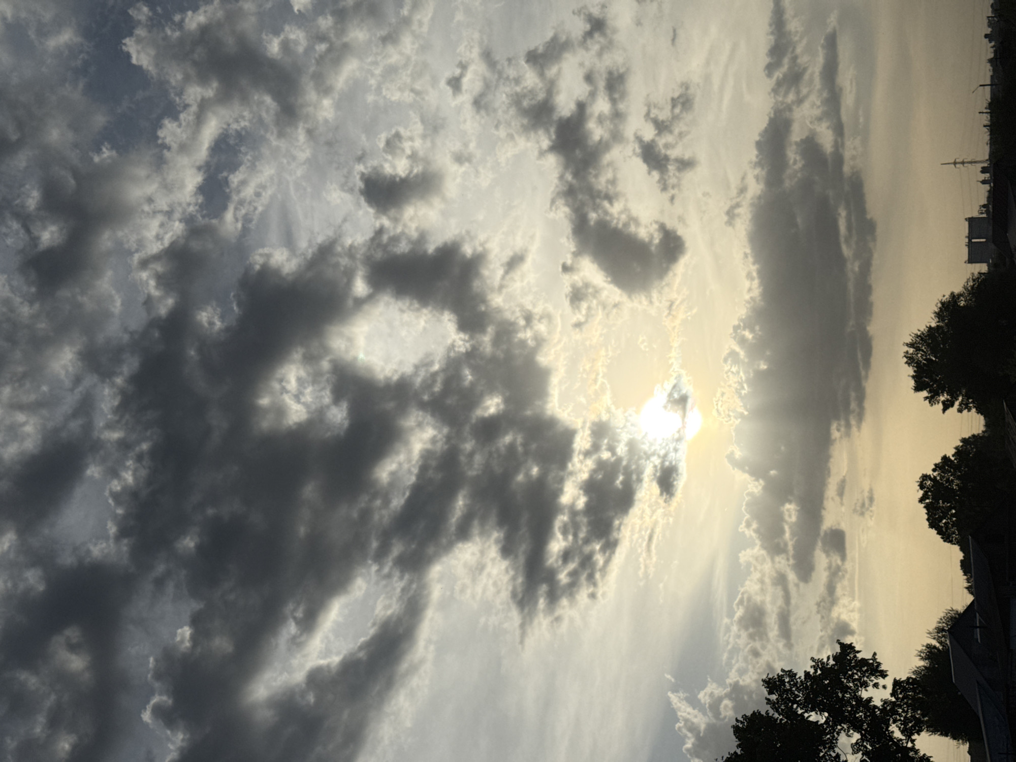 The image depicts a sky filled with clouds, with the sun visible through a gap in the cloud cover. The lower right portion of the image shows silhouettes of trees and rooftops. In the far distance along the horizon, utility poles and buildings are faintly visible. The sky transitions from lighter, brighter areas around the sun to darker, more opaque cloud formations. The location is not confirmed to be Ленино, Russia and is based on the assumption that the photograph was taken there.