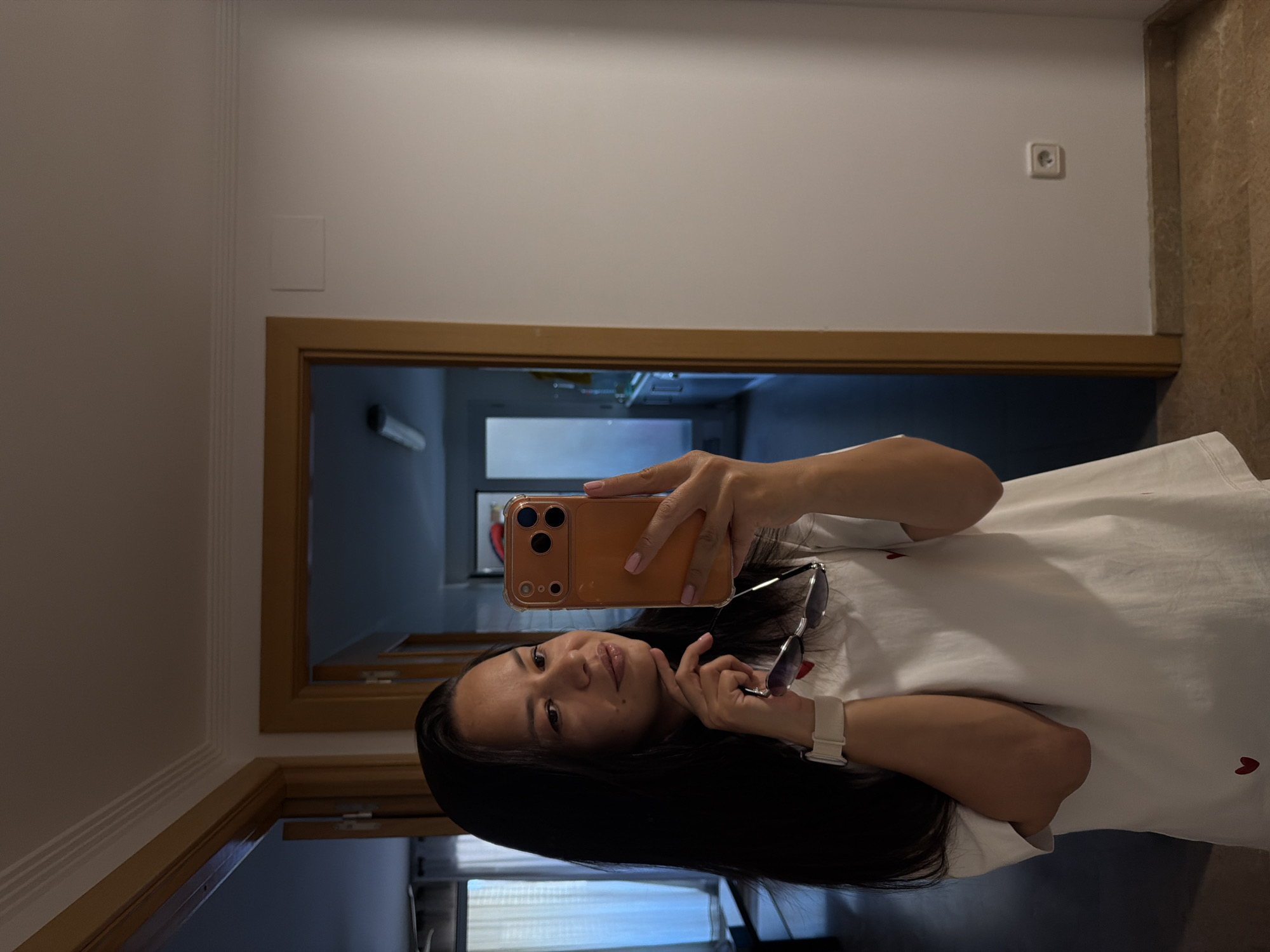 A woman with long, dark hair takes a selfie in a mirror. She is wearing a white t-shirt with small red hearts printed on it and a white smartwatch on her left wrist. She is holding a pair of sunglasses in her right hand and resting her chin on her left hand. The background appears to be an interior hallway with doorways leading to other rooms. The lighting suggests it is daytime.