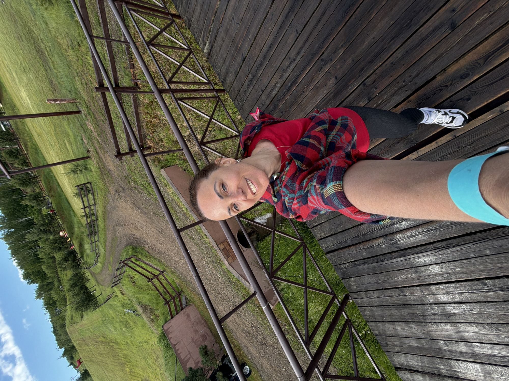 A woman with brown hair, wearing a red shirt, a red and blue plaid shirt, black leggings, and white athletic shoes, is taking a selfie on a wooden platform with a metal railing. She is smiling and looking at the camera. Behind her, a green, hilly landscape is visible, with a dirt road winding up the hill and trees at the top. The location appears to be an outdoor recreational area, possibly a park or a resort, in Birobidzhan, Russia, given the context. The sky is partly cloudy and blue.