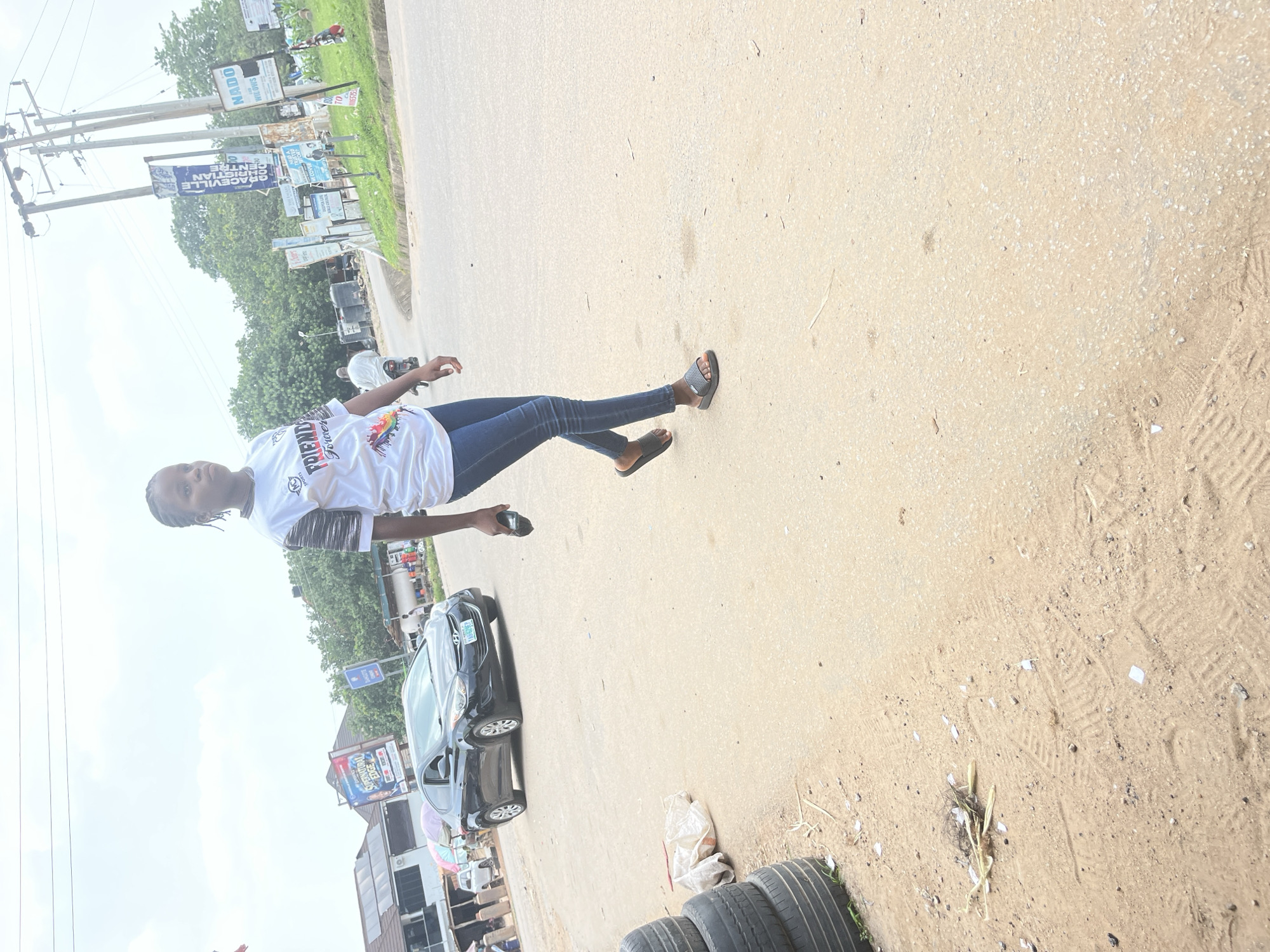 A young woman walks on a paved road in Abuja, Nigeria. She is wearing a white t-shirt with graphics and blue jeans, and black sandals. She is holding a black object in her right hand. In the background, there are trees, buildings, and utility poles. A black car is parked on the side of the road. Several tires are stacked on the left side of the image. The road is dusty and there is some debris on the ground. The sky is overcast.