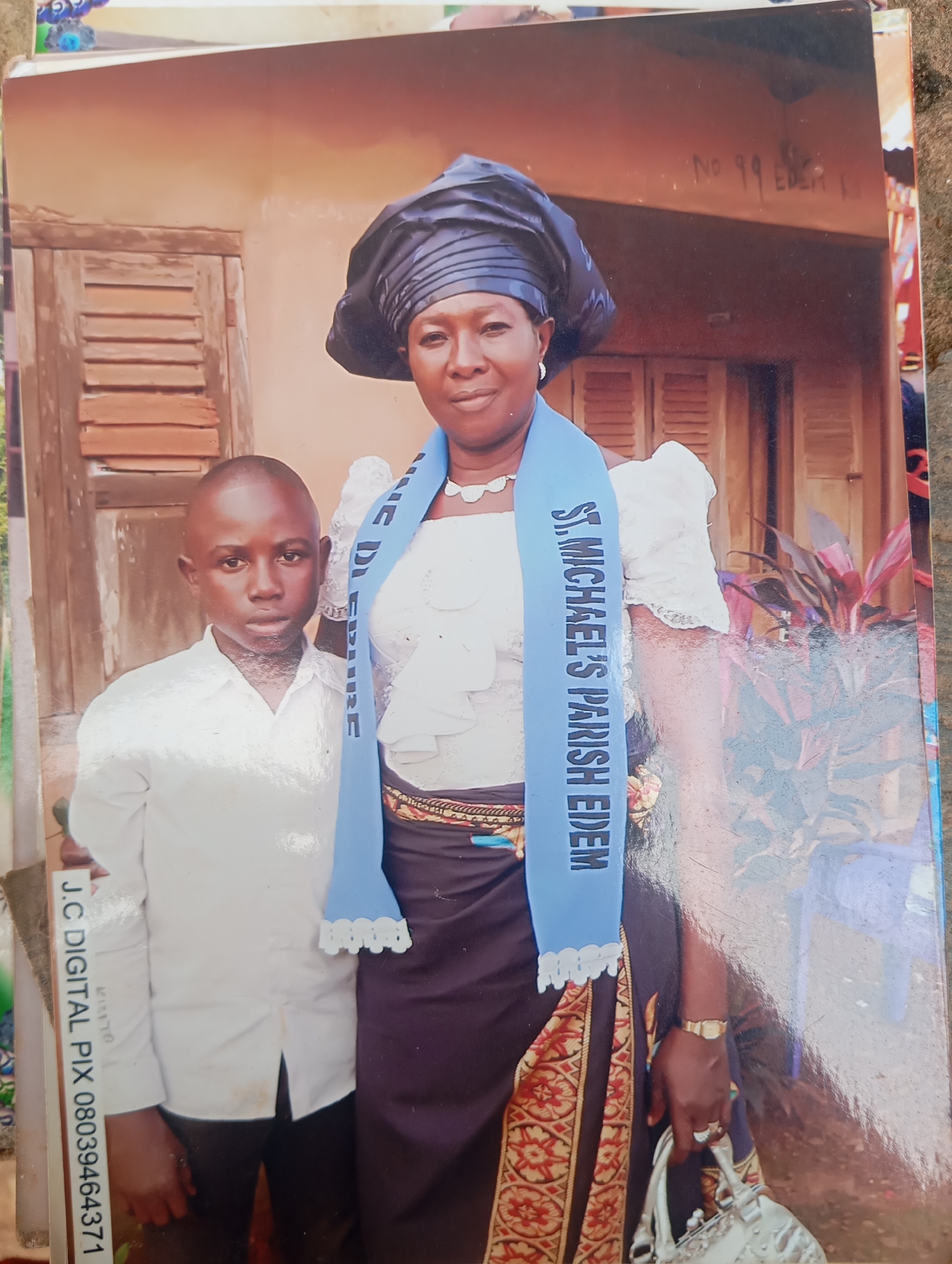 A medium shot photograph captures a woman and a boy standing in front of a building in Dutsen Alhaji, Nigeria. The woman, on the right, is wearing a black headwrap, a white ruffled blouse, and a dark skirt with a patterned trim. A blue sash draped around her neck reads "ST. MICHAEL'S PARISH EDEM." She has a watch on her left wrist and rings on her fingers, and is holding a silver purse. The boy, on the left, is dressed in a white collared shirt and dark pants. He has a serious expression. The background shows a weathered wooden door with louvers, part of a building wall, and some foliage. A handwritten sign above the woman's head reads "NO 99 EDEN 1". The bottom left of the image features a watermark that says "J.C DIGITAL PIX 08039464371".