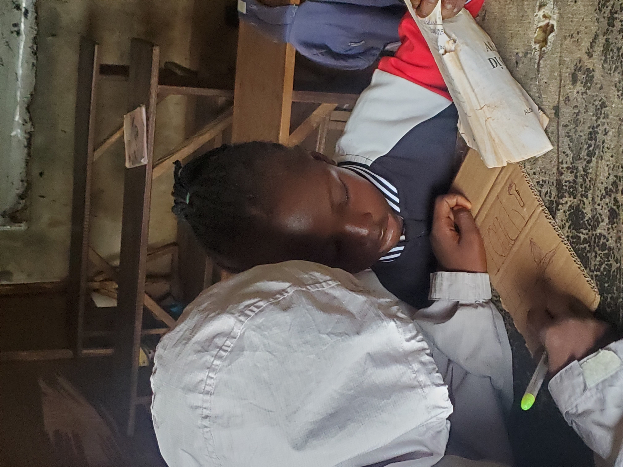 A photograph shows a child with braided hair, wearing a dark top with white stripes at the neck, lying on what appears to be a table. The child's eyes are closed. A person wearing a light-colored jacket is sitting next to the child, holding a piece of cardboard on which something is drawn. They are holding a pen with a green tip. A piece of paper is near the cardboard. The table's surface looks worn. The background shows shelves and what appears to be the interior of a building. The location is specified as the city of Ukwu-Abwa, Nigeria.