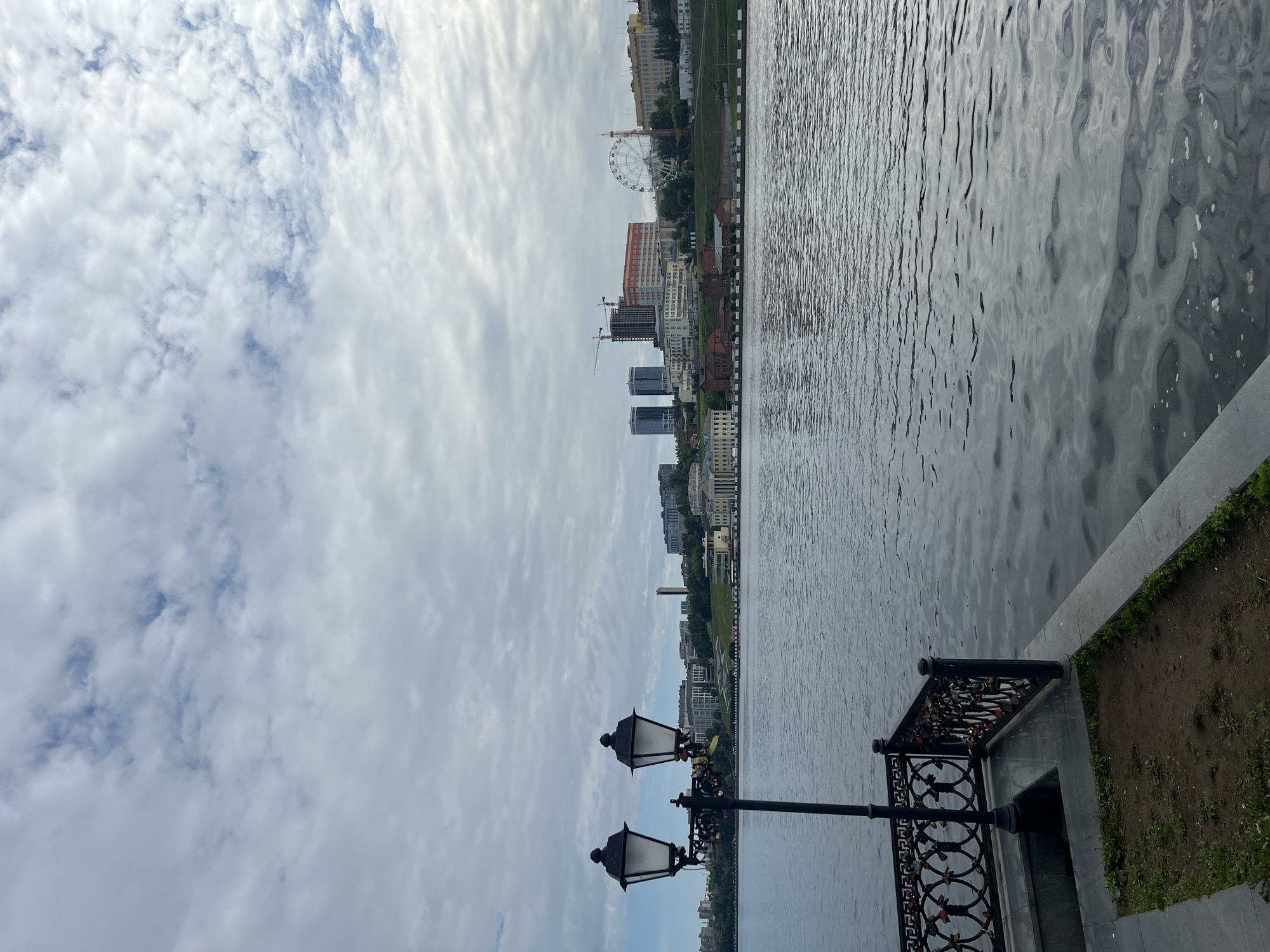 This image depicts a wide view of the Izhevsk Pond embankment in Izhevsk, Russia, on an overcast day. In the immediate foreground, a black ornamental lamppost with two traditional lanterns stands on a gray paved surface. Numerous "love locks" are attached to the base of the lamppost and to the black decorative metal railing running along the edge of the embankment. A small, sparse patch of dirt and grass is visible next to the paved area.
The central and lower portions of the image are dominated by the Izhevsk Pond, characterized by ripples on its dark, reflective surface, which mirrors the muted gray tones of the sky. No boats or individuals are present on the water.
Across the pond, the distant bank rises, showcasing a cityscape. On the right side of the far bank, a prominent white Ferris wheel is visible. To its left and extending across the bank are various multi-story buildings, encompassing both older architectural styles, including a structure with columns on the far left, and modern constructions. Several visible construction cranes indicate ongoing development. Green foliage is scattered across the hillside and between buildings.
The sky overhead is predominantly covered with dense, white and light-gray stratocumulus clouds, with intermittent small patches of faint blue sky visible through breaks. The overall lighting is diffuse, indicative of an overcast daytime setting. No individuals are discernible within the scene.