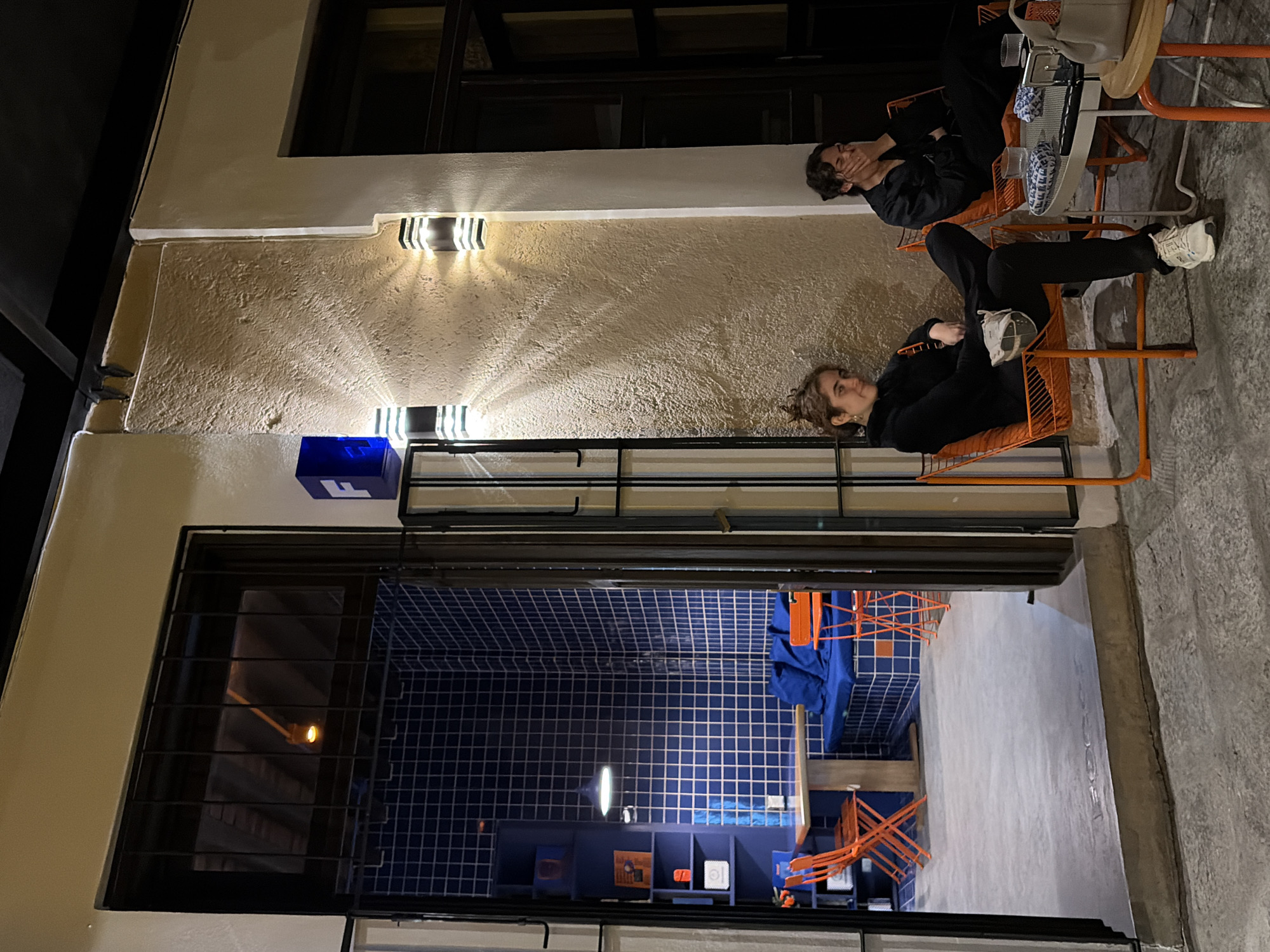 A photograph shows two individuals in an outdoor setting in Puebla, Mexico. The two people are seated in bright orange, wire-frame chairs around a small, round table. The woman closest to the frame has her legs stretched out, and is looking at the camera. The man is behind her and is in a darker hoodie. There is a beige building facade in the background, illuminated by built-in lights on the exterior. The interior of the building is visible through a window behind iron bars, displaying a blue-tiled space with orange accents.