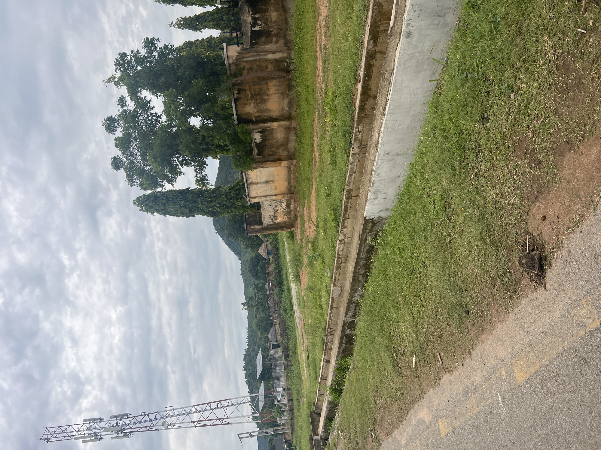 The image depicts an outdoor scene in Kona Garu, Nigeria, under a cloudy sky. The primary subjects are a concrete drainage channel, a grassy area, and a road with yellow markings. In the background, there are several structures including old, weathered buildings and tall, slender trees. Further in the distance, a lush green hillside with scattered structures can be seen. On the left side of the image, a large metal tower-like structure, possibly a communication tower or part of a construction crane, is visible against the sky. The overall setting appears to be a semi-urban or rural area. There is no clear activity or event taking place, and no people are visible. The time of day is indeterminate due to the overcast weather, but the lighting suggests it is daytime. The environment appears somewhat neglected, with peeling paint on the buildings and overgrown grass.