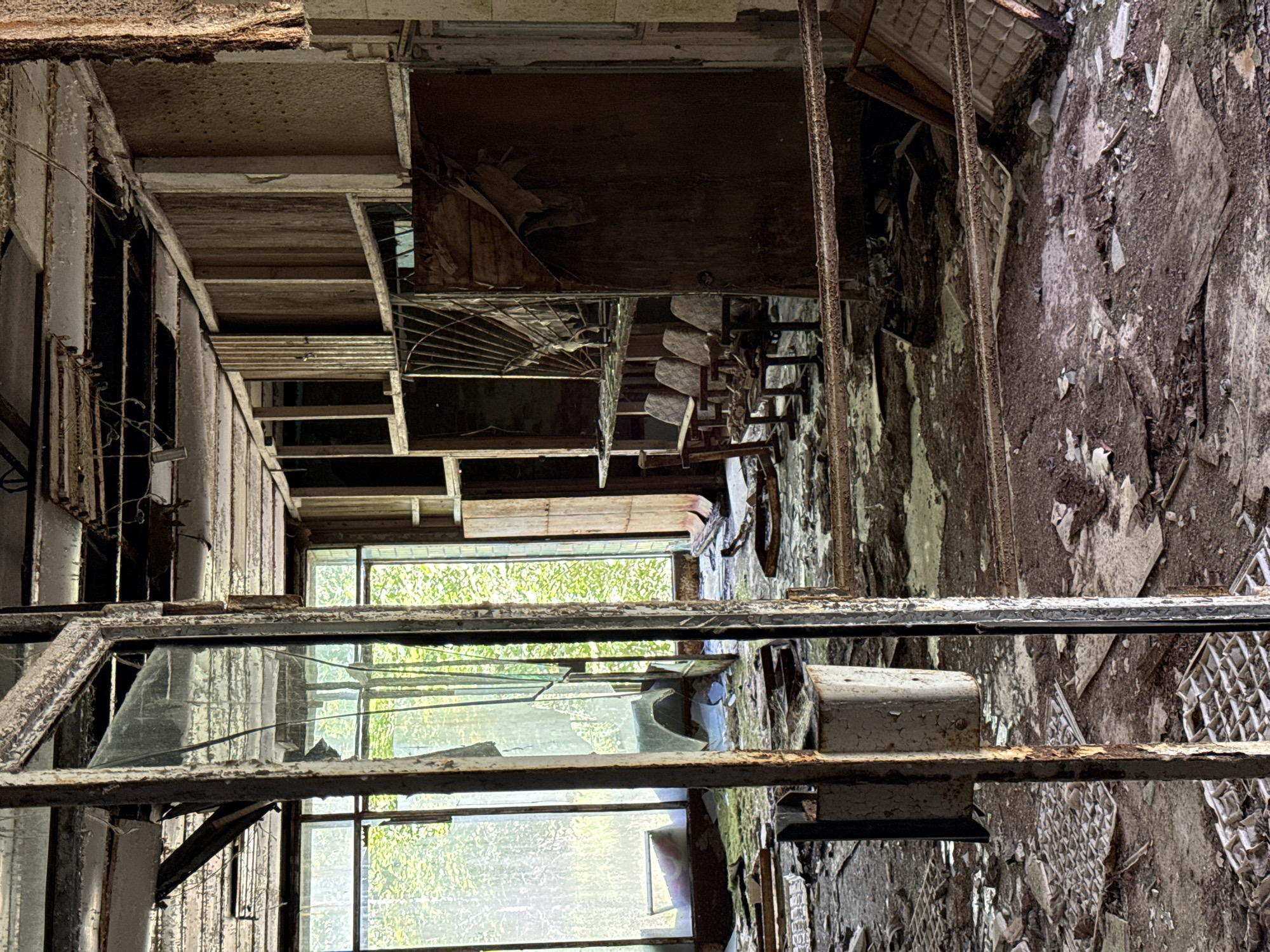 The interior of an abandoned building in Prypiat, Ukraine is captured in this image. The walls are heavily damaged, with crumbling plaster and exposed brick. A series of broken windows face outward, offering glimpses of vegetation beyond. Rusted metal frames and debris are scattered throughout the scene. The space is filled with decaying materials and evidence of disrepair.