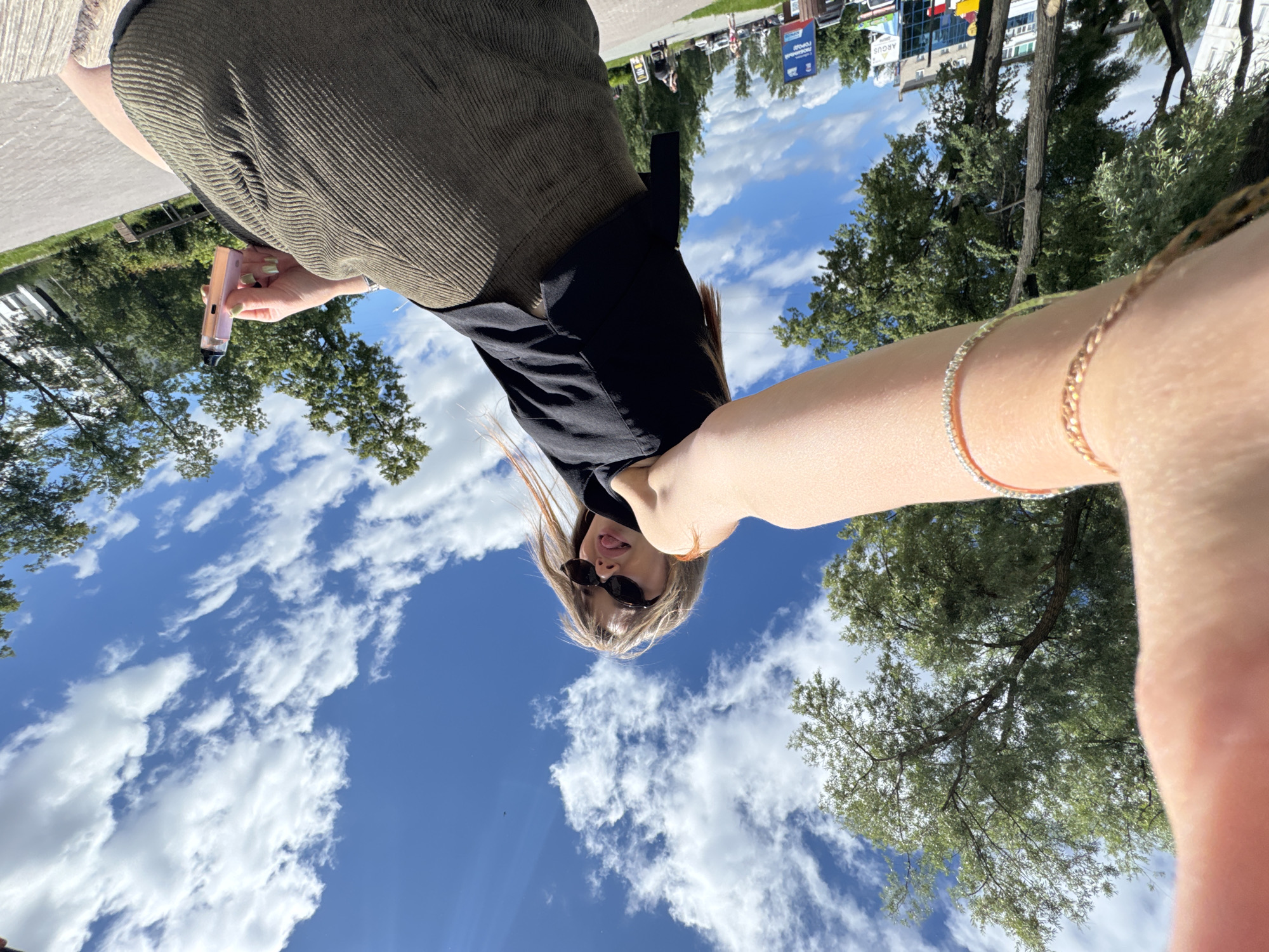 The image is a selfie taken outdoors with a wide-angle lens, showing a young woman with blonde hair, sunglasses, and a black top paired with a ribbed olive green skirt. She is holding a pink vape device in her right hand. The background features a bright blue sky with fluffy white clouds, lush green trees, and a paved walkway. In the distance, buildings and street signs are visible, including one with "ARGUS" on it and another with Russian text that translates to "Chaikovsky City". The perspective suggests the woman is sitting or leaning back.
