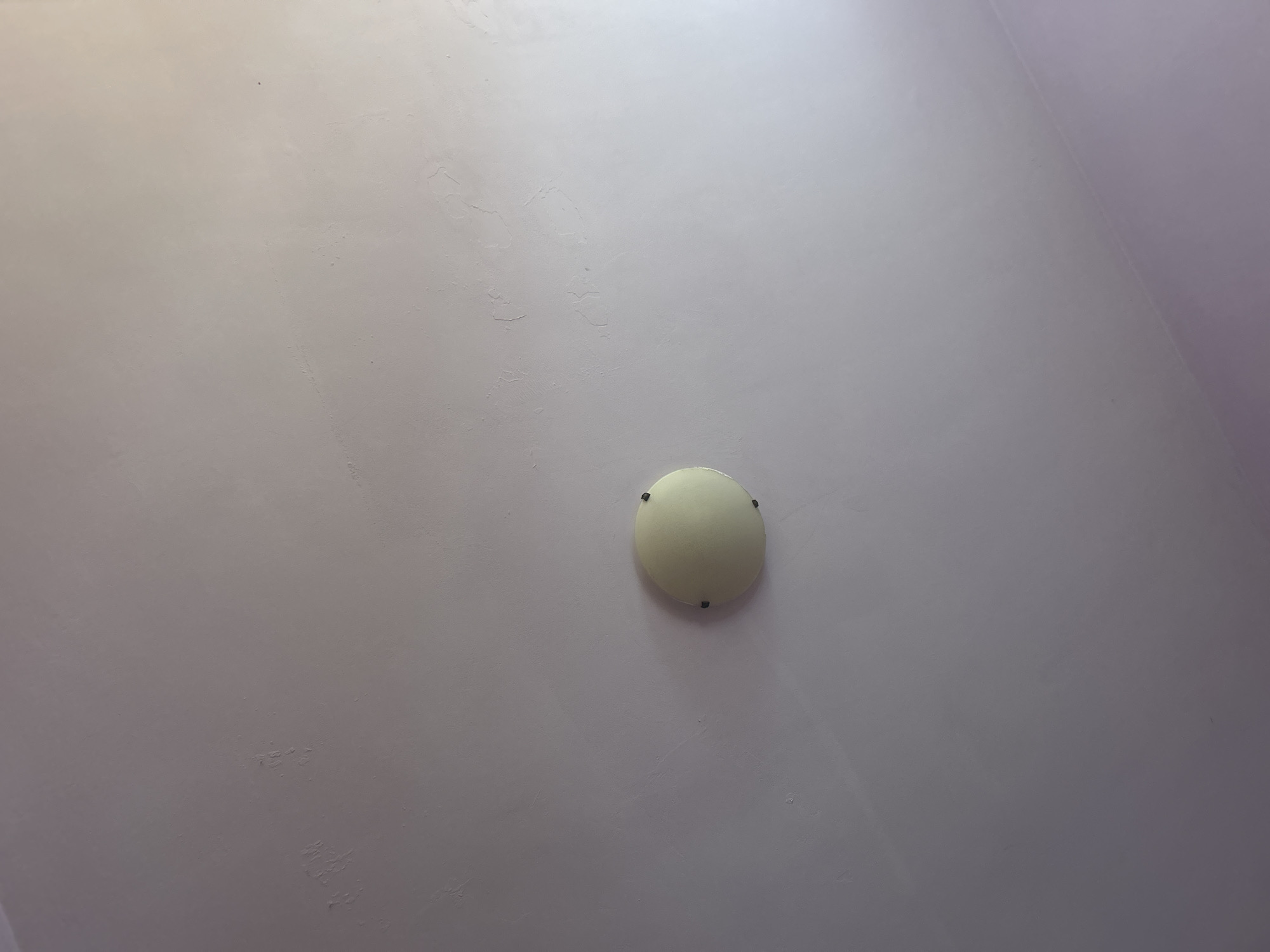 The image shows a close-up of a light fixture on a ceiling. The fixture is circular and appears to be made of a frosted glass or plastic material, with visible dark screws securing it to the ceiling. The ceiling itself is painted a light color and shows some imperfections and texture. The lighting in the image is somewhat uneven, with a brighter area on the right side, suggesting a light source outside the frame. The context provided, mentioning Plovdiv, Bulgaria, is not visually discernible from the image itself. There are no people, actions, or interactions depicted.