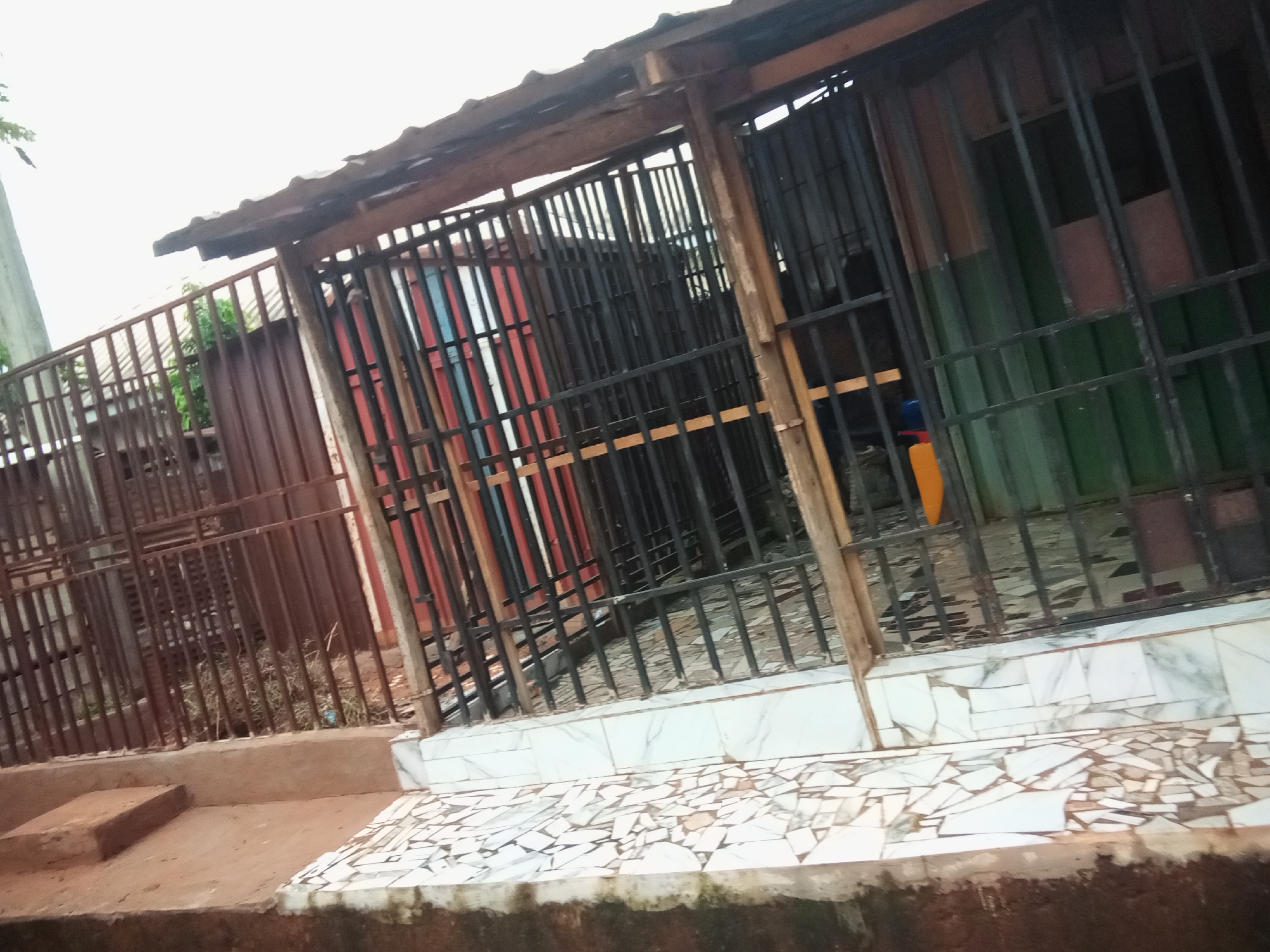 The image shows a section of a building with a barred window and a corrugated iron fence in Oko Town, Nigeria. The building appears to be a shop or dwelling, with a rough wooden frame supporting a corrugated iron roof. The barred window has black metal bars and behind it, there are vertical corrugated iron sheets of various colors, including red and green. The fence is made of rusted corrugated iron sheets. In front of the building, there is a concrete step and a tiled floor made of broken white and brown tiles. The area appears to be outdoors, with dry grass and some debris visible behind the fence. The sky is overcast, suggesting a cloudy day.