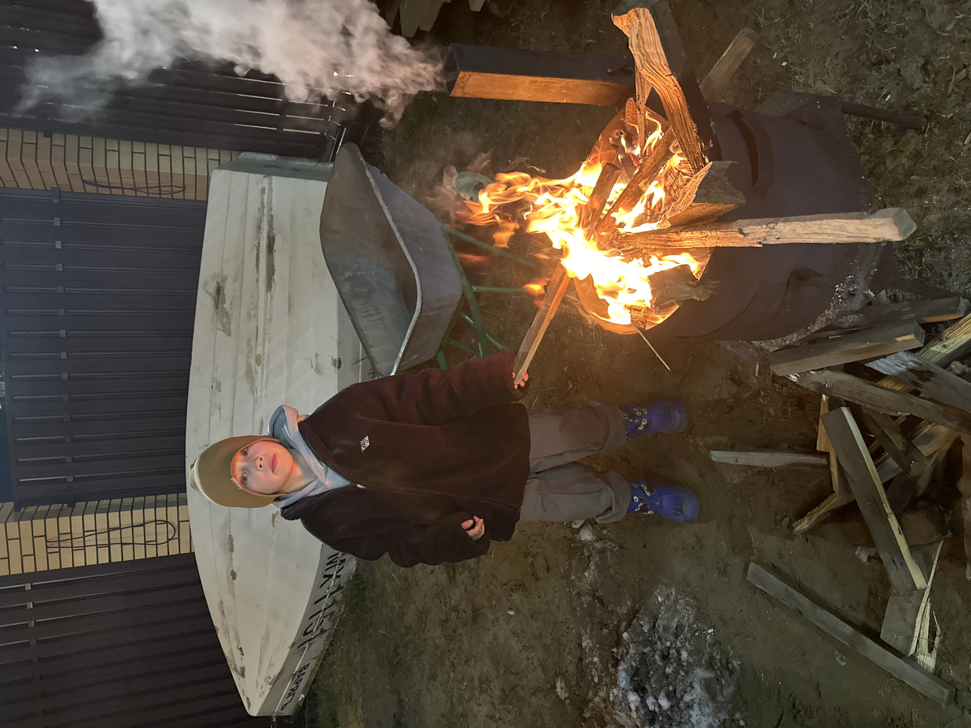 A person stands in a dirt-covered yard, next to a burning fire in a black drum. The individual wears a brown jacket, gray pants, a hat, and blue boots. Wood is stacked on the ground near the fire. Behind the person, a white boat rests on its side, with visible lettering in Russian. A wheelbarrow sits next to the boat. A fence and brick wall are visible in the background. Smoke rises from the fire. Patches of snow or ice are scattered on the ground. The location is слобода Старая, Russia.