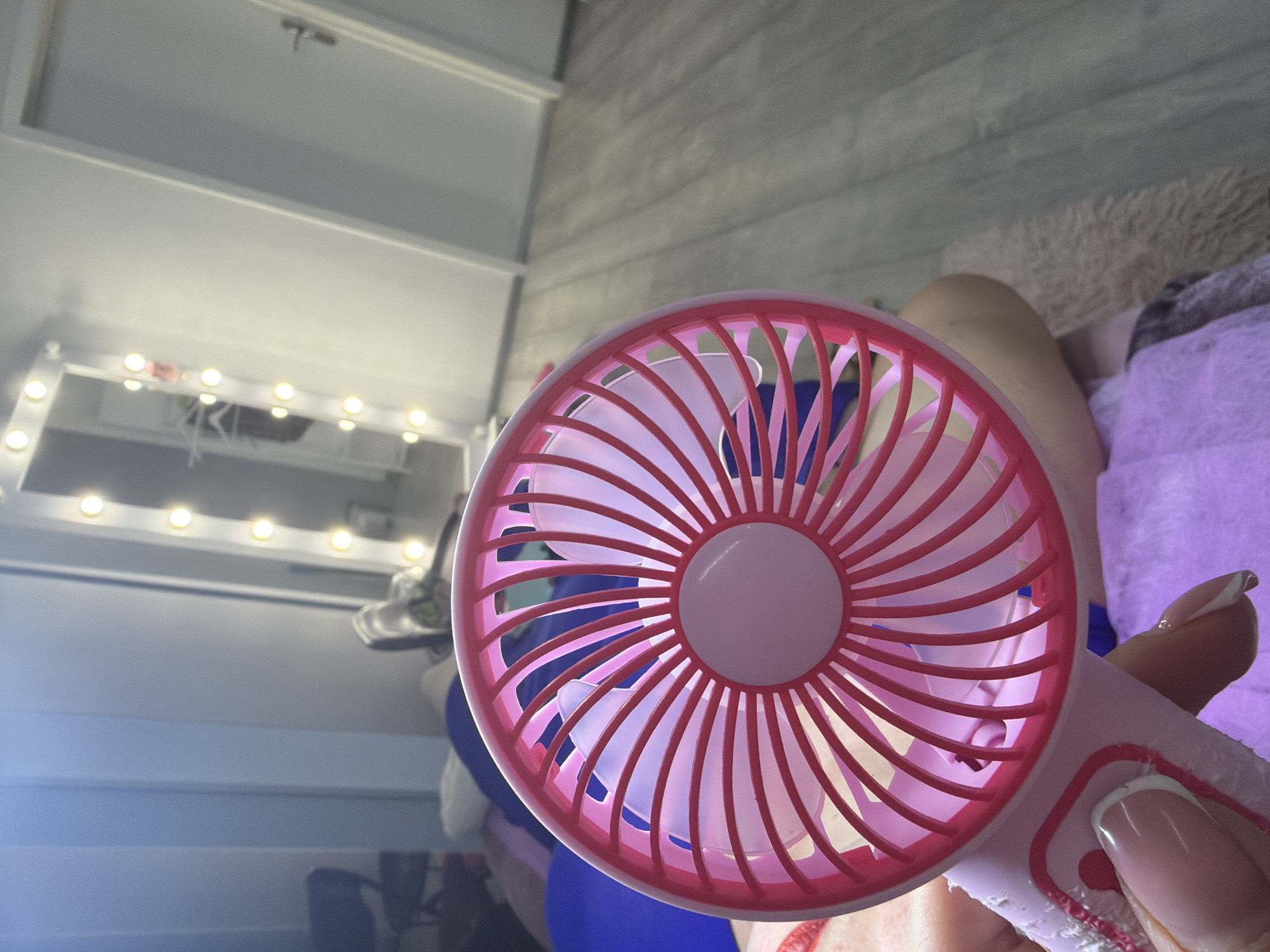A person holds a pink and white handheld fan with a red grill in a room. The fan is positioned in the foreground, with its blades visible through the grill. The person's hand, with manicured nails, is holding the fan's handle. In the background, a blurred reflection of a vanity mirror with lights is visible. The room appears to be a bedroom or dressing area, with a wooden floor and a door on the right. The overall impression is of a personal, indoor setting.