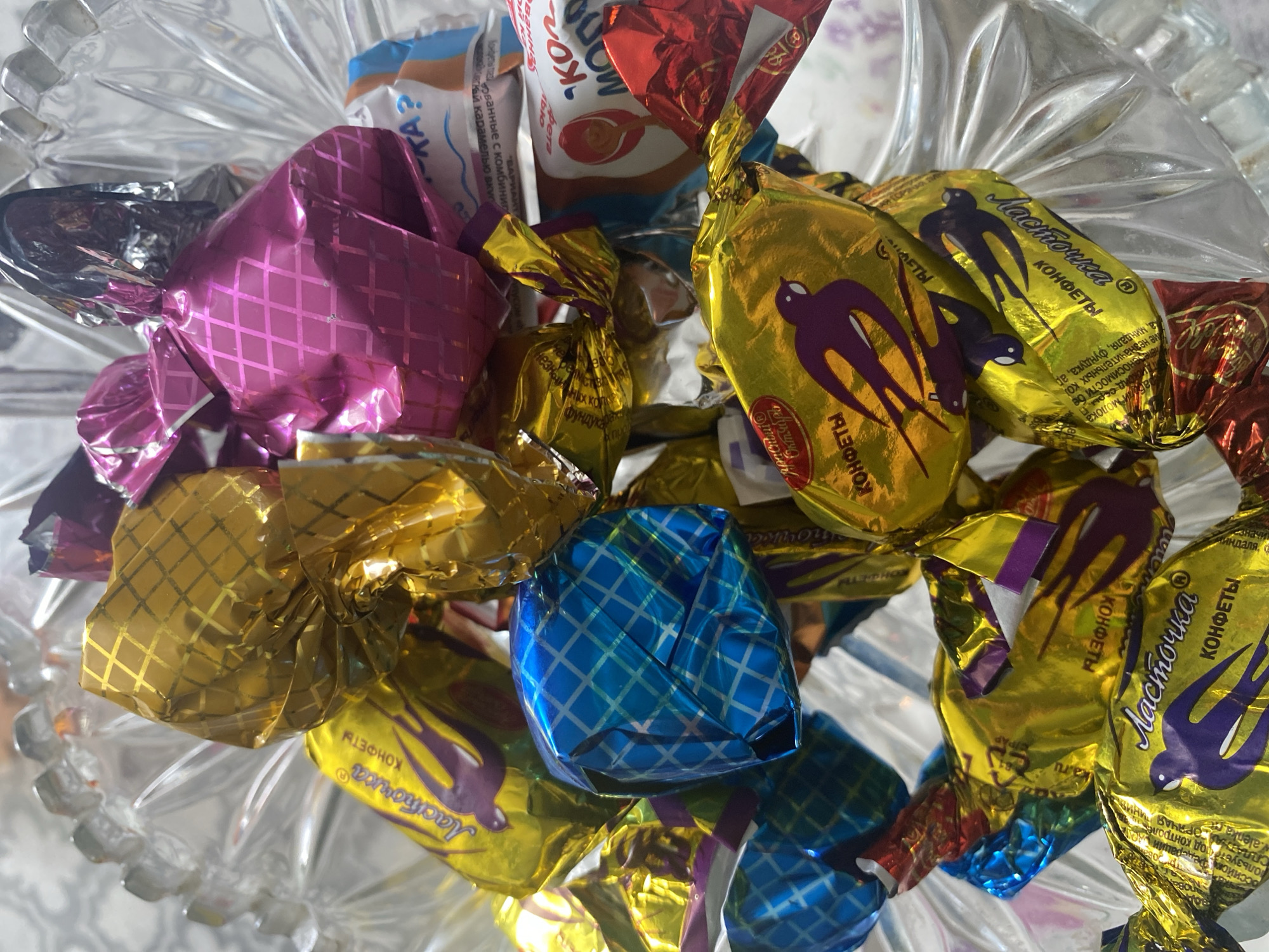 A close-up, overhead shot displays a glass bowl filled with an assortment of individually wrapped candies. The candies are in various colorful foil wrappers, including gold, pink, blue, and red. Some of the gold wrappers feature an image of a purple bird and the text "Ласточка" (Lastochka) and "КОНФЕТЫ" (konfety), indicating they are "Swallow" brand candies. Other wrappers have geometric patterns. The bowl itself appears to be made of clear, patterned glass. The background is out of focus, showing a light-colored surface with a subtle pattern. There is no indication of the city of Chontaul, Russia, in the image.