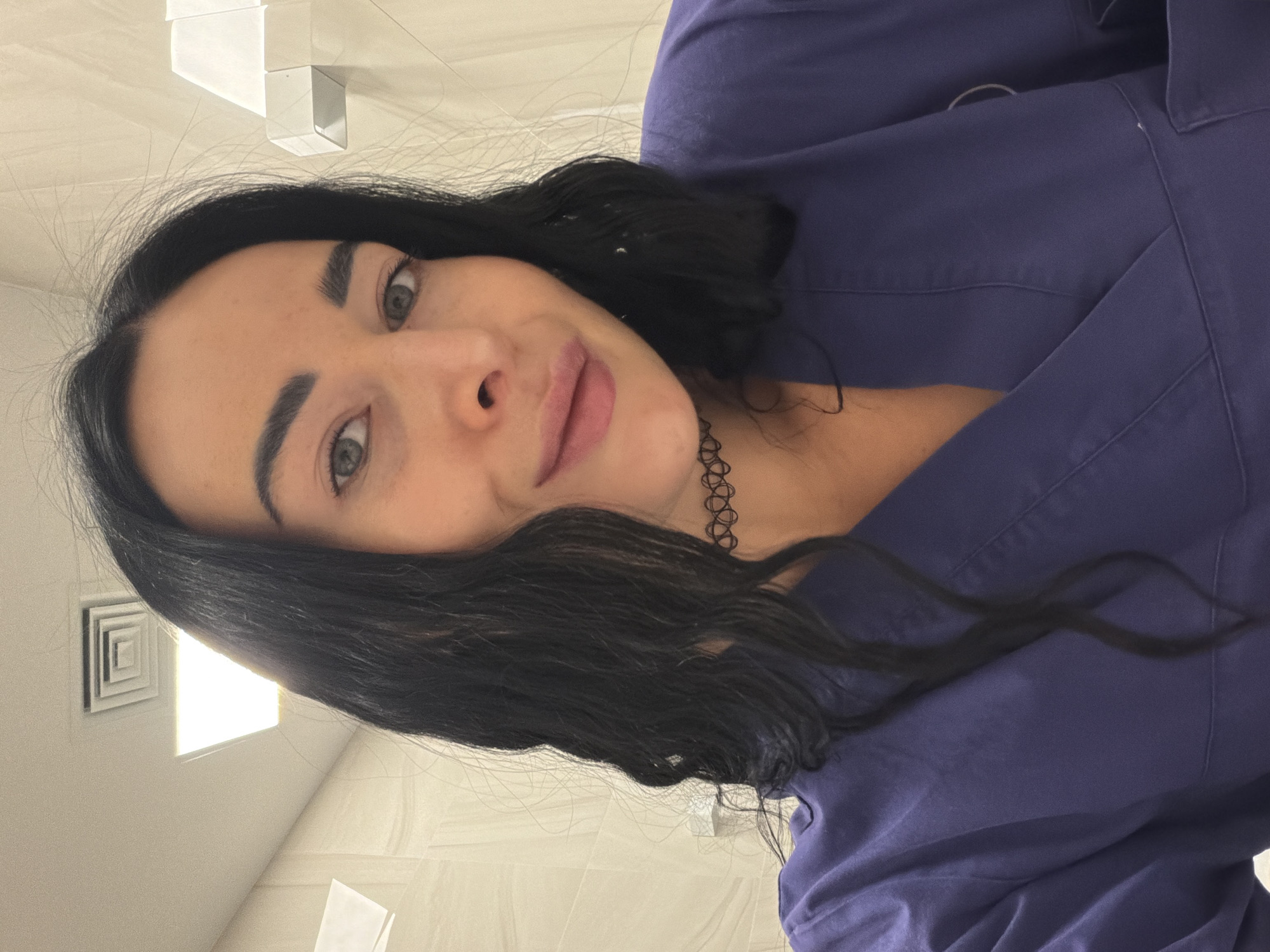 A young woman with dark hair, dark eyebrows, and light eyes is posing for a selfie. She is wearing a dark blue collared shirt and a black choker necklace. The background is a bright, tiled wall, with a ventilation duct visible in the upper left corner. The location cannot be determined to be Saint Petersburg, Russia.