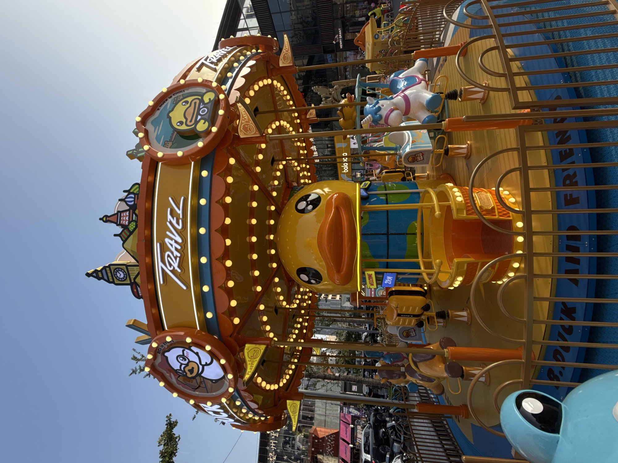 The image shows a colorful B. Duck themed carousel in Yiwu, China. The carousel features various duck-shaped and other cartoon characters as rideable figures, illuminated by numerous small yellow lights. The top of the carousel is adorned with depictions of iconic landmarks such as Big Ben, suggesting a "travel" theme. In the background, there are modern buildings, shops, and some parked vehicles and motorbikes, indicating an urban setting. A few people are visible in the distance. The sky is clear and blue, and the overall lighting suggests it is daytime, likely late afternoon due to the warm tones. The B. Duck logo and the words "TRAVEL" are prominently displayed on the carousel. The ground surrounding the carousel has a blue textured surface with the words "B. DUCK AND FRIENDS" written on it. A blue, anthropomorphic character is visible in the foreground, partially obscuring the carousel's fence.