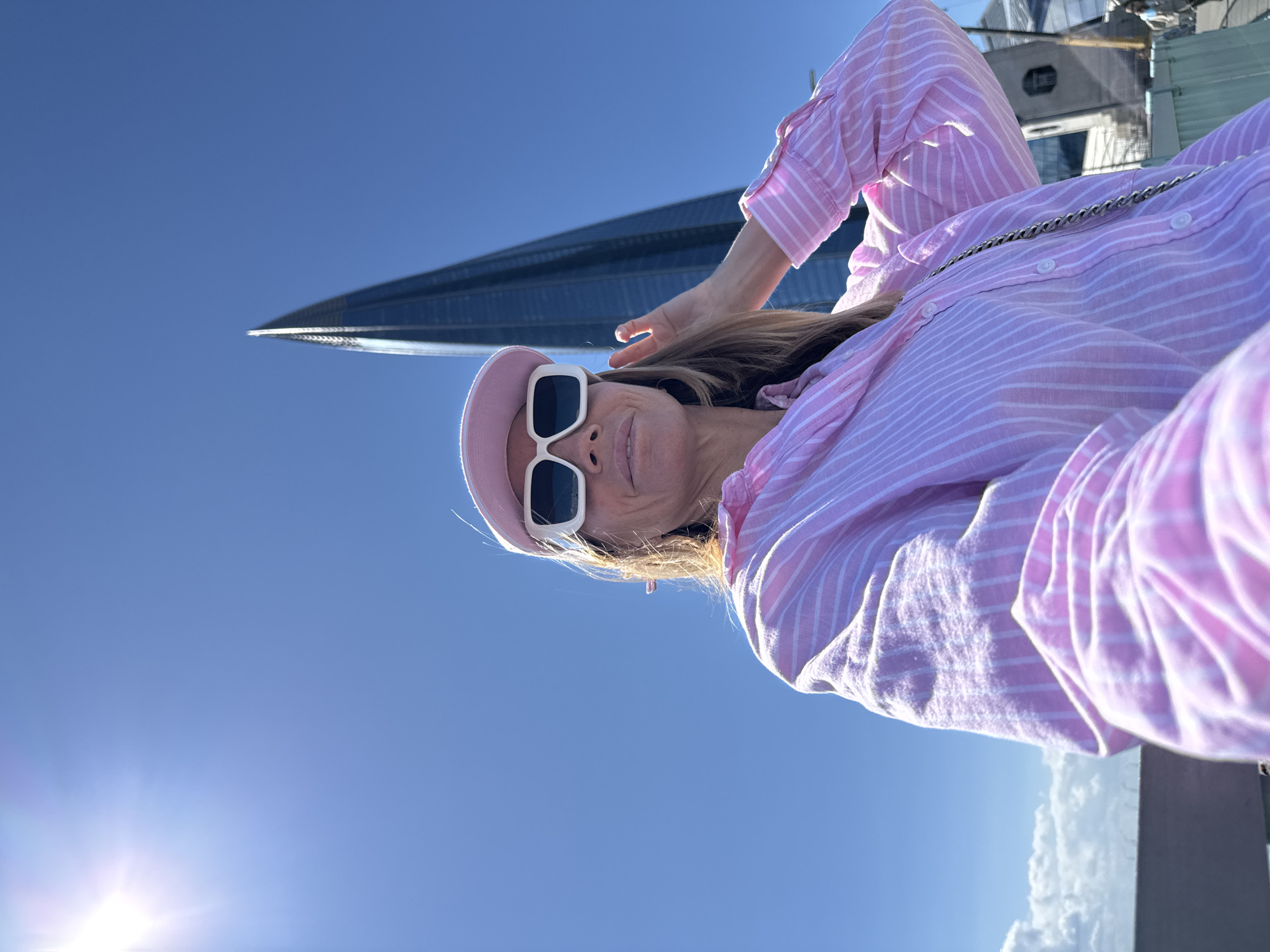 An image, rotated 90 degrees clockwise (appears horizontal), features a woman posing outdoors against a clear blue sky, with a prominent modern skyscraper in the background. The woman, positioned on the right, wears a light pink and white striped long-sleeved button-down shirt, a pink visor, and white square-framed sunglasses. A silver chain, likely a bag strap, is visible across her chest. She has light brown hair and a subtle, relaxed smile, looking directly at the camera. Her right arm is raised with her hand near her head in a casual gesture.
Behind her, on the left side of the frame, stands the sharp, pointed spire of a tall, dark glass skyscraper. This distinctive architecture strongly resembles the Lakhta Center in Saint Petersburg, Russia, which aligns with the provided location context. The sky is a brilliant, cloudless blue, save for some wispy white clouds visible towards the lower right. Bright sunlight causes a noticeable lens flare in the lower left corner of the sky, indicating a clear, sunny day, likely mid-day or early afternoon. No visible text is present in the media file. The overall scene conveys a feeling of relaxation and enjoyment in a modern urban setting.