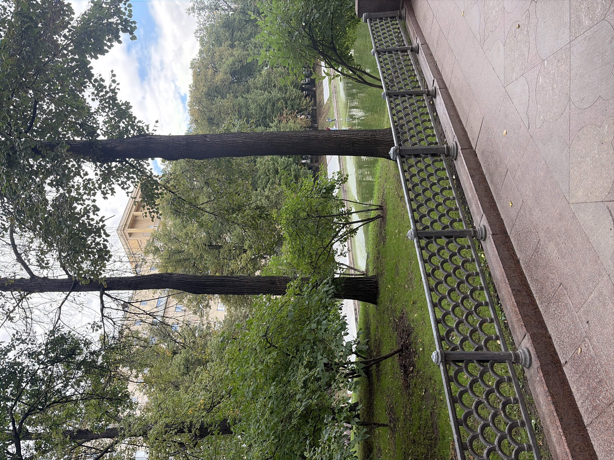 A view of a park in Moscow, Russia, shows a pond bordered by trees and a building in the background. A pathway made of pink granite tiles leads to the right of the frame, and a decorative iron fence separates the path from a grassy area and the pond. The pond's surface is green and reflects the surrounding trees. Several people are visible near the pond, some appearing to be fishing. The building in the background is light yellow and has multiple windows. The sky is cloudy with patches of blue. The scene suggests a pleasant day in a city park.