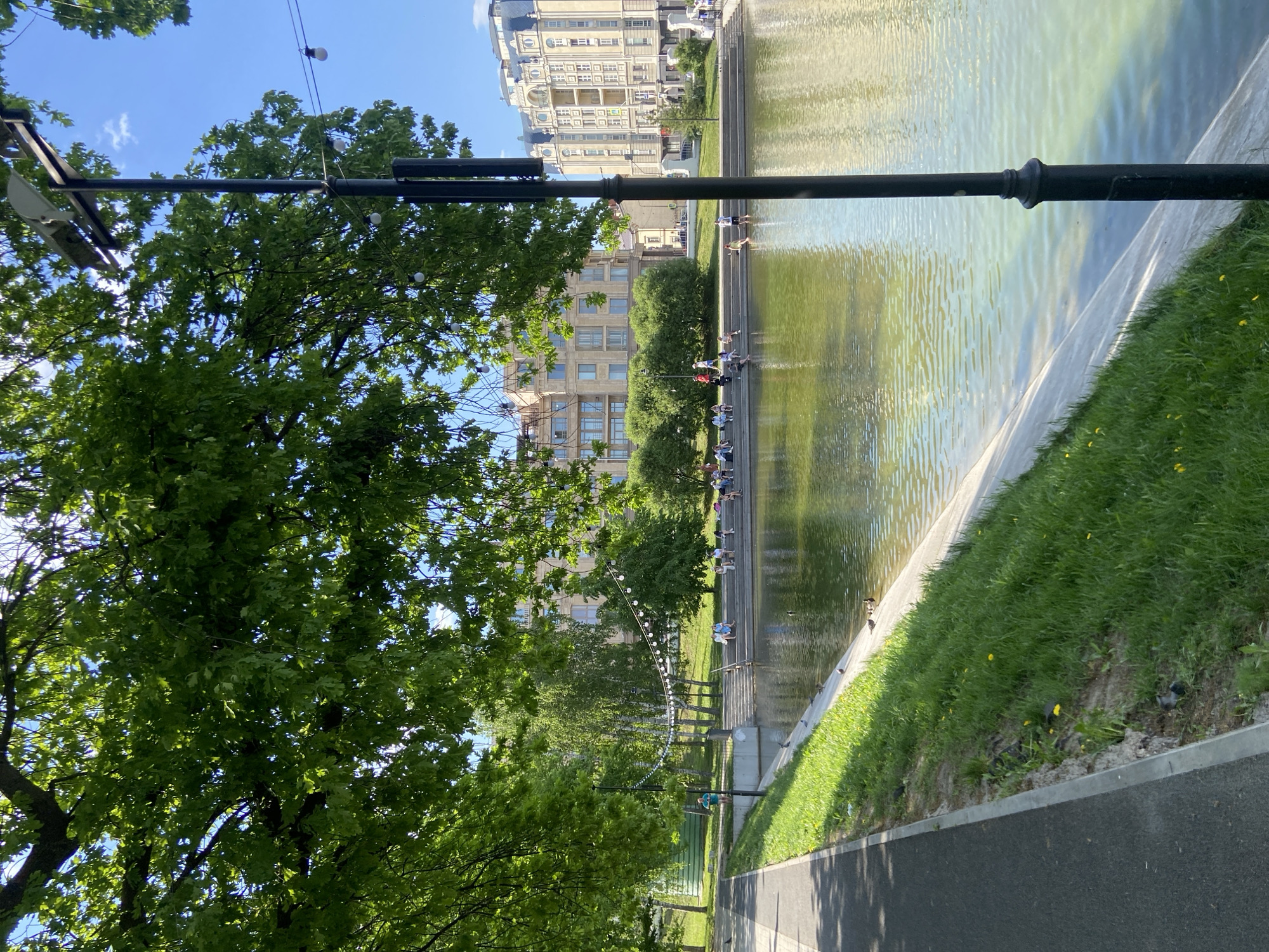 The image shows a park scene in Kazan, Russia. A body of water sits to the right, bordered by concrete and grass. People are sitting on steps leading into the water. Buildings can be seen in the background. A tree and a lamp post stand to the left of the image. A paved path is in the foreground.