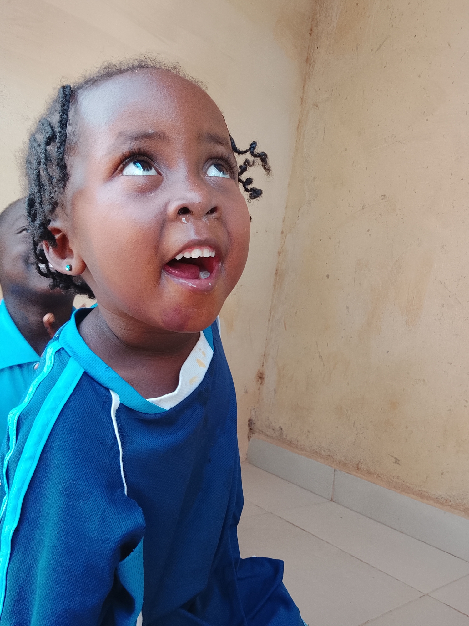 A young child with dark skin and braided hair, wearing a blue athletic shirt with white trim around the collar and light blue trim on the shoulders, is looking upwards with an open mouth. A second child is partially visible behind the first, also wearing a blue shirt. The background is a tan wall with white tile flooring. This image was taken in Kuru Dungus, Nigeria.