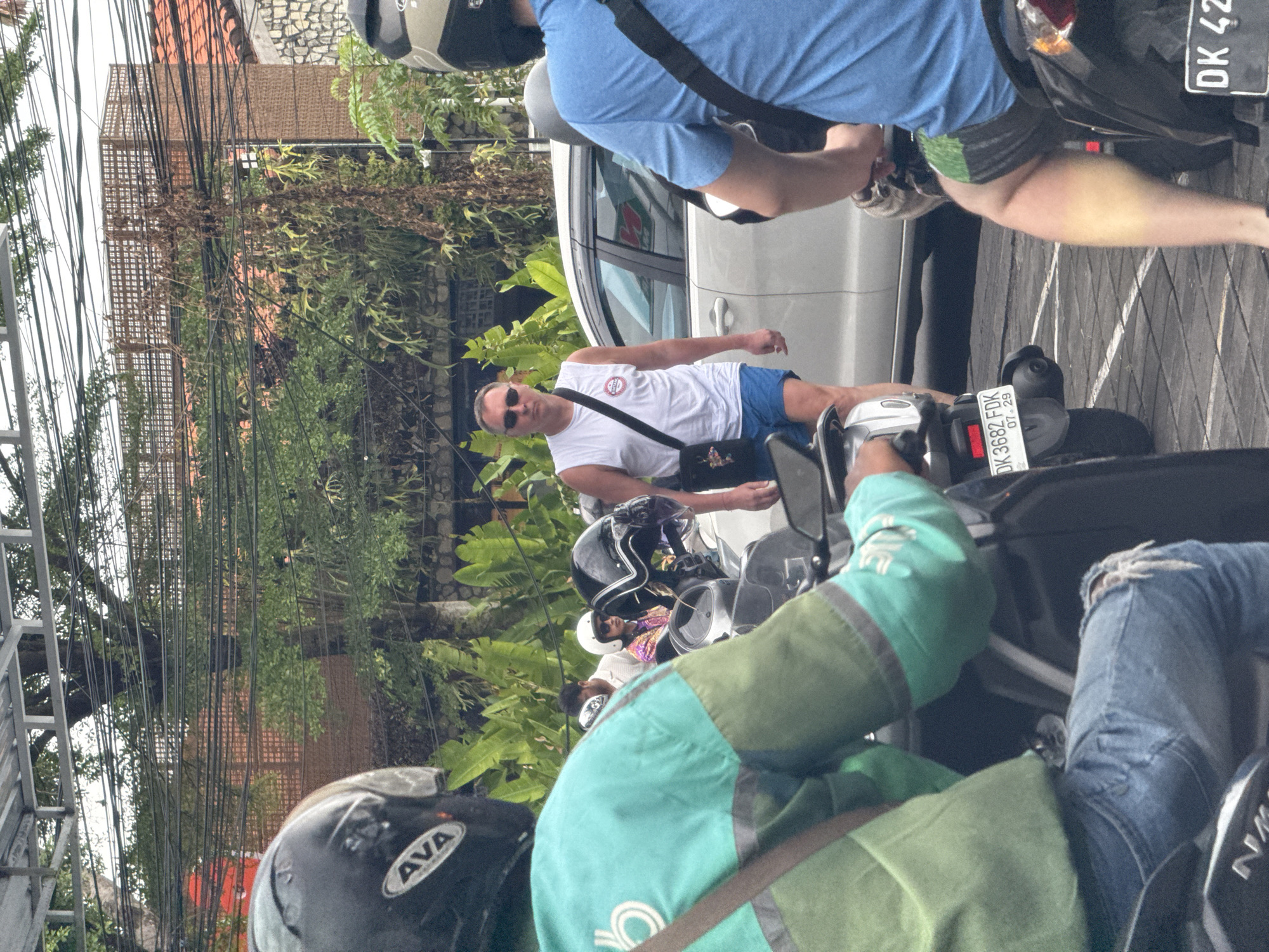 The image captures a bustling daytime street scene in North Kuta, Indonesia, predominantly featuring people on scooters and a white car. The perspective is from behind a foreground scooter rider, looking forward into traffic.
In the immediate foreground, the back of a person wearing a turquoise and green jacket, possibly a ride-share driver, and blue ripped jeans is visible, seated on a scooter. A black helmet with the white "AVA" logo rests on another scooter to their left.
Further into the scene, a man stands next to a scooter, wearing a white t-shirt with a red and white circular logo, blue shorts, and sunglasses. He carries a black shoulder bag. Behind him, a white car is parked or slowly moving. Another person on a scooter, wearing a light blue t-shirt and dark shorts, is partially visible.
Visible license plates on the scooters include "DK 3682 FDK 07.29" and a partial "DK 42". "DK" indicates the province of Bali, aligning with the North Kuta location.
The background is rich with tropical foliage, including trees and dense bushes, suggesting a lush environment. Overhead, numerous black power and communication lines crisscross the frame, a common sight in urban areas of Southeast Asia. Parts of buildings with reddish-brown tiled roofs and textured walls can be seen through the greenery.
The scene appears to be a typical moment of urban traffic, likely with people commuting or navigating the local streets during daylight hours. No specific event or strong emotions are discernible.