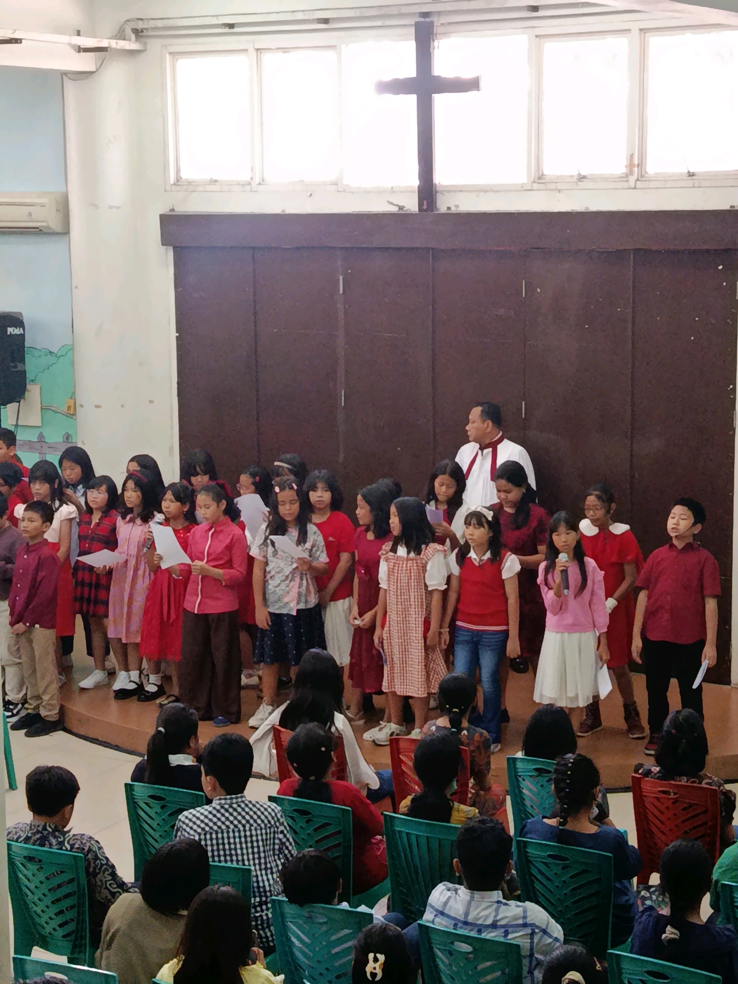 In East Jakarta, Indonesia, a group of approximately 25 children and one adult male stand before a wooden wall with three large windows behind them, illuminated by bright light. A large wooden cross is affixed to the wall above the windows. The children, appearing to be of elementary school age, are gathered on a raised platform, some holding papers, possibly reading lyrics or a script. A young girl in the front row holds a microphone. The adult male, wearing a white shirt with a red collar, stands slightly behind the children, appearing to lead or oversee them. The children are dressed in a variety of clothing, many in shades of red and pink, suggesting a performance or religious gathering. In the foreground, several rows of empty green plastic chairs are visible, some with people seated, their backs to the camera. The overall environment suggests an indoor space, likely a church or community hall.