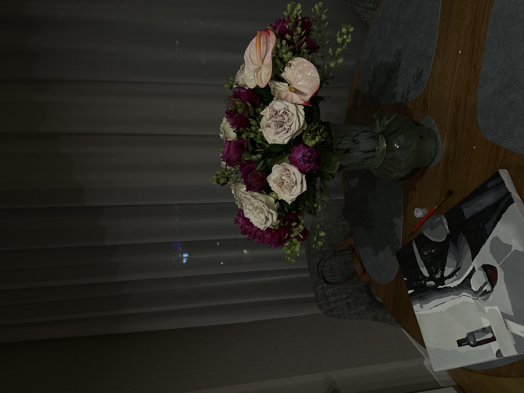 A bouquet of flowers sits in a vase on a wooden table, next to a canvas painting. The bouquet contains roses, purple flowers, and two pink anthuriums. The vase is green and marbled. The painting is black and white, and features a bottle. The table is covered in placemats. A chair is visible next to the table. Behind the bouquet is a window with closed blinds. The location is not discernible from the image.