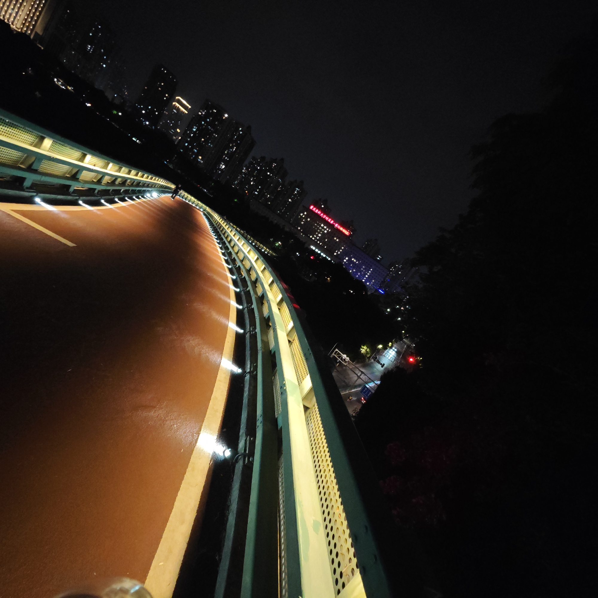 The image depicts an elevated pedestrian walkway or bridge at night in the Yuzhong District of Chongqing, China. The walkway has a reddish-brown surface with yellow lane markers and is illuminated by recessed lights along the side. A green railing borders the walkway. In the background, there are city buildings with lit windows, some of which have illuminated red Chinese characters on the roof. A dark area, likely trees or vegetation, is visible to the right of the walkway. A street intersection with traffic lights is seen below the walkway on the right. The image is taken at an angle, giving a skewed perspective of the scene.