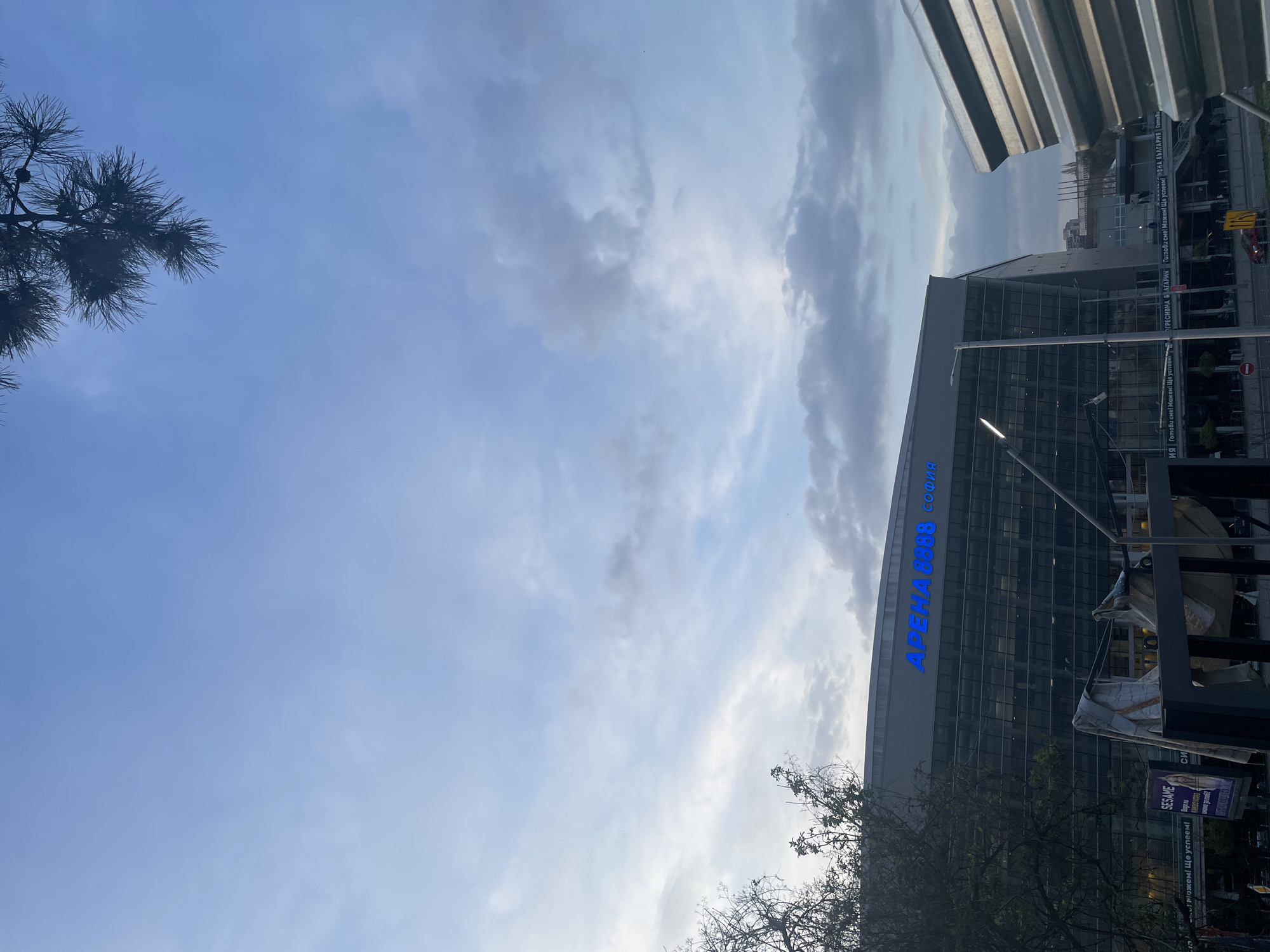 An outdoor, daytime shot captures a modern urban landscape in Sofia, Bulgaria, dominated by a large building on the right under a partly cloudy sky. The building is a multi-story structure with a glass facade, reflecting the surroundings. Prominently displayed on its upper section is a large blue sign with the Cyrillic text "АРЕНА 8888 СОФИЯ" (ARENA 8888 SOFIA).
The sky, occupying a significant portion of the left and upper frame, shows patches of clear blue interspersed with white and grey clouds, suggesting a dynamic weather condition, possibly late afternoon or early evening given the diffused light. A dark, pine-like tree branch with needles extends from the upper left corner into the clear blue sky. In the lower right foreground, the bare branches of another tree are visible, partially obscuring the building's base.
Additional urban elements include a corrugated metal structure partially visible at the very top right, and what appear to be streetlights or poles. Several banners or posters are attached to structures near the building. Visible text on these includes "SESAME" and the repeated phrase "Готови сме! Можем! Ще успеем!" (We are ready! We can! We will succeed!), accompanied by variations of "АГРЕСИВНА БЪЛГАРИЯ" (Aggressive Bulgaria), likely a promotional or political slogan. The scene is devoid of visible people, conveying a calm, architectural focus.