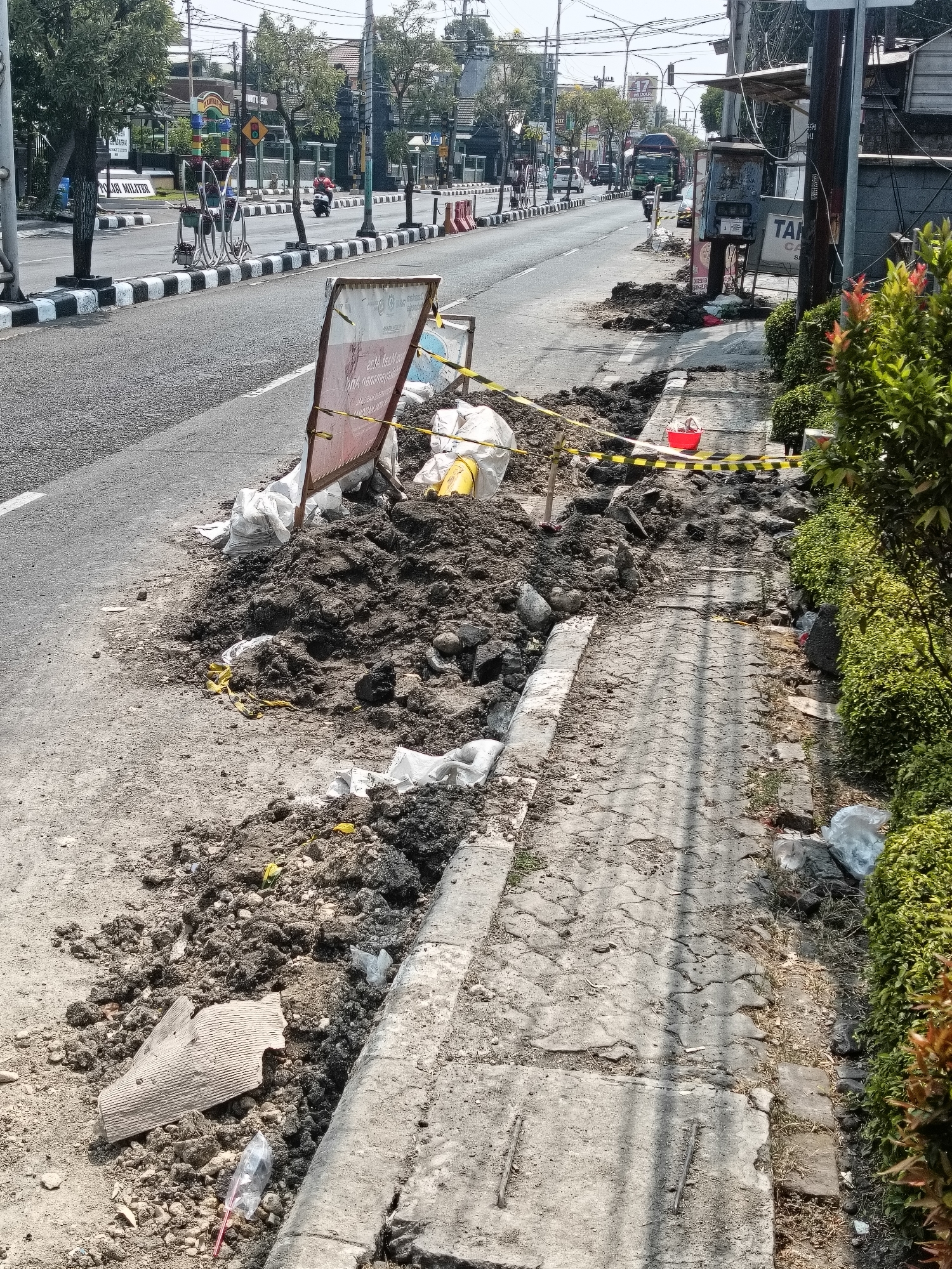 The image shows a section of a street in Gapurosukolilo, Indonesia, where construction or repair work is underway. Piles of excavated dirt and debris are present along the sidewalk and encroaching onto the road. Yellow and black caution tape is used to cordon off the work area. Several white sacks, likely containing construction materials or disposed of waste, are visible. A red bucket is also present near the sidewalk. The road itself appears to be asphalt, with white lane markings. In the background, a busy street with vehicles, trees, and buildings can be seen. The overall impression is one of ongoing public works disrupting the urban environment.