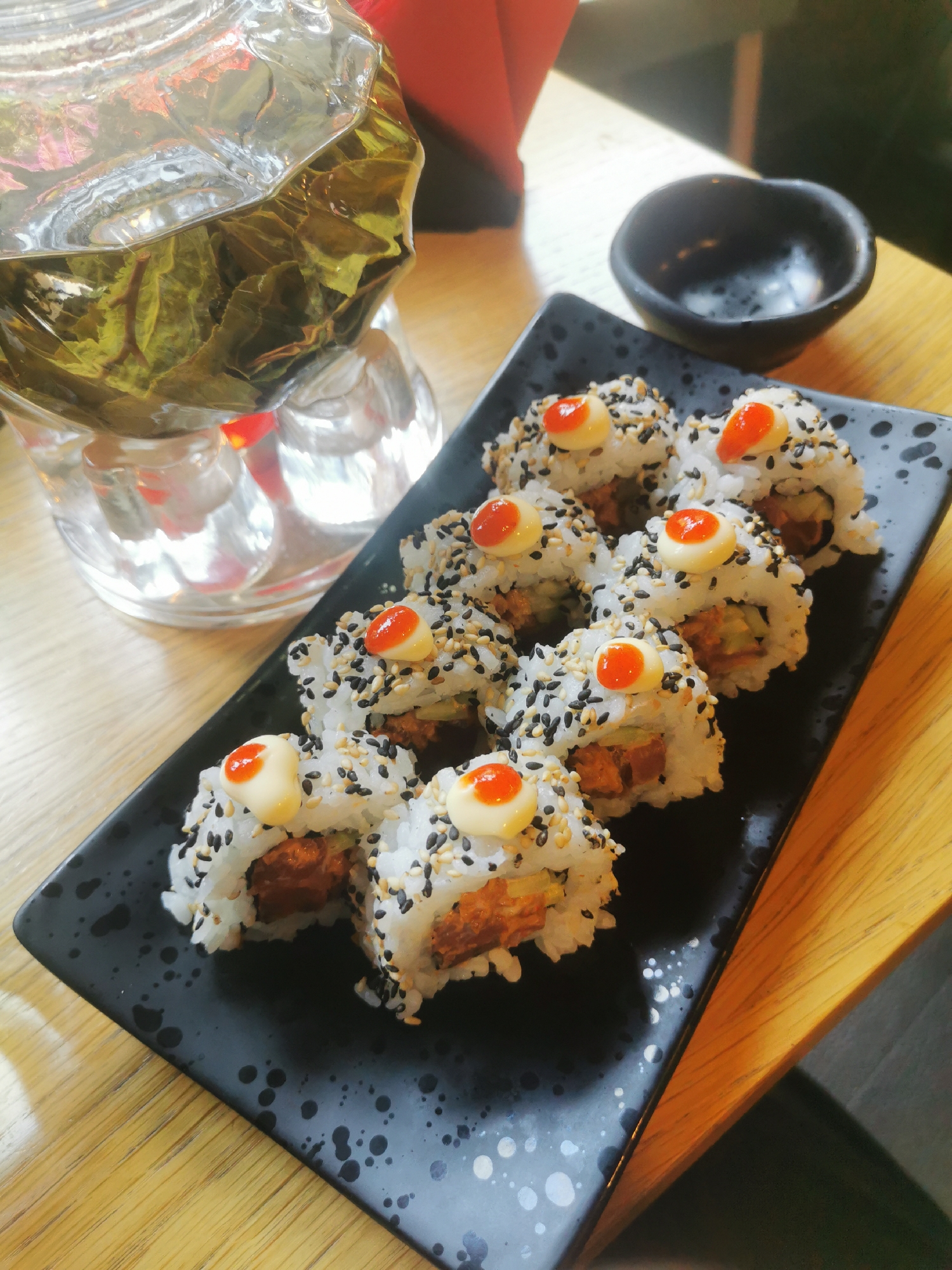 The image captures a meal served on a light-colored wooden table, likely within a restaurant or cafe in Blagoevgrad, Bulgaria. The primary subject is a black rectangular plate presenting eight generously sized sushi rolls. These rolls, possibly Uramaki, are made with white rice and liberally coated with a mix of black and white sesame seeds. Each roll is topped with a dollop of creamy yellowish-white sauce, accented with a small, vibrant dot of orange or red sauce. The visible filling within the rolls appears to be a cooked, reddish-brown ingredient, perhaps fried chicken or tempura, combined with some green elements like cucumber.
To the upper left of the sushi plate, a clear glass teapot or carafe is prominently featured, filled with water and numerous steeping green tea leaves, suggesting hot tea is being served. Reflections of a red or orange object are visible through the base of the teapot. A small, dark, unglazed ceramic bowl, presumably for soy sauce or dipping, rests behind and to the right of the sushi plate.
The wooden table surface has a visible grain, and in the background, a darker surface, possibly another table or part of the restaurant's decor, can be seen, along with a faint red hue. The lighting indicates an indoor setting. There is no visible text in the image. The scene conveys a pleasant and appetizing dining experience.