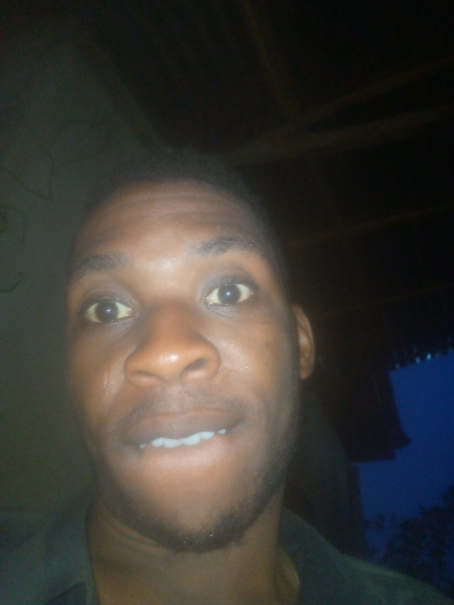 The image is a close-up portrait of an adult individual, likely male, with dark skin, looking directly at the camera. His eyes are wide open, and his mouth is slightly agape, revealing his lower teeth, which suggests an expression of surprise, curiosity, or being caught off guard. There is a visible sheen or glint on his skin, particularly on his forehead, nose, and cheeks, possibly due to sweat or reflections from the lighting. He has some stubble or short facial hair on his chin and jawline.
The setting appears to be dimly lit, likely at night or in a very dark room, with the subject's face illuminated by a bright, direct light source, possibly a camera flash. The background is mostly dark and underexposed. Vague, dark shapes in the upper background suggest indoor structures, possibly wooden beams or a ceiling. In the lower right portion of the image, there is a distinct patch of dark blue, hinting at an outdoor night sky or a distant element seen through an opening. The subject is wearing dark-colored clothing, visible around his neck and shoulders. No discernible objects besides the person are clearly visible. There is no visible text.