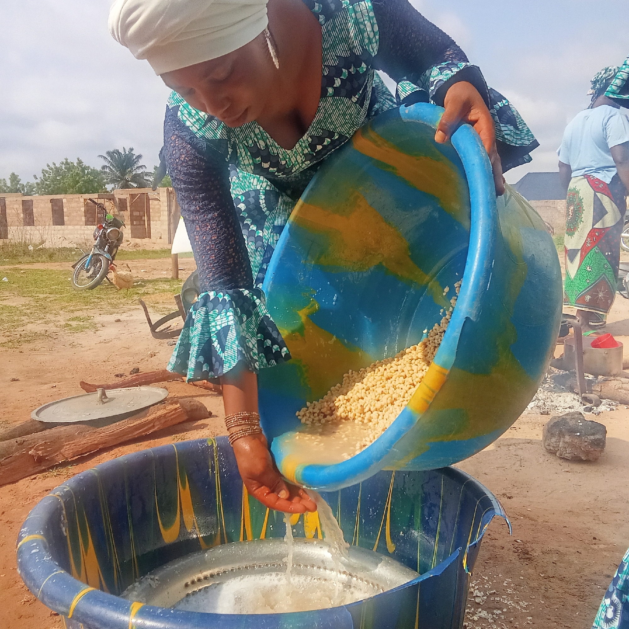 In Doma, Nigeria, a woman wearing a patterned blue and green dress and a white head covering is pouring a tub of tan, round, shelled beans into another blue tub containing water with bubbles. She is wearing multiple gold bracelets on her left wrist. In the background, there is a motorcycle, a partially built brick building, a woman in a blue shirt and patterned skirt, firewood, a metal pot, and burning embers. The ground is red dirt. The sky is partially obscured by the woman and the tub.