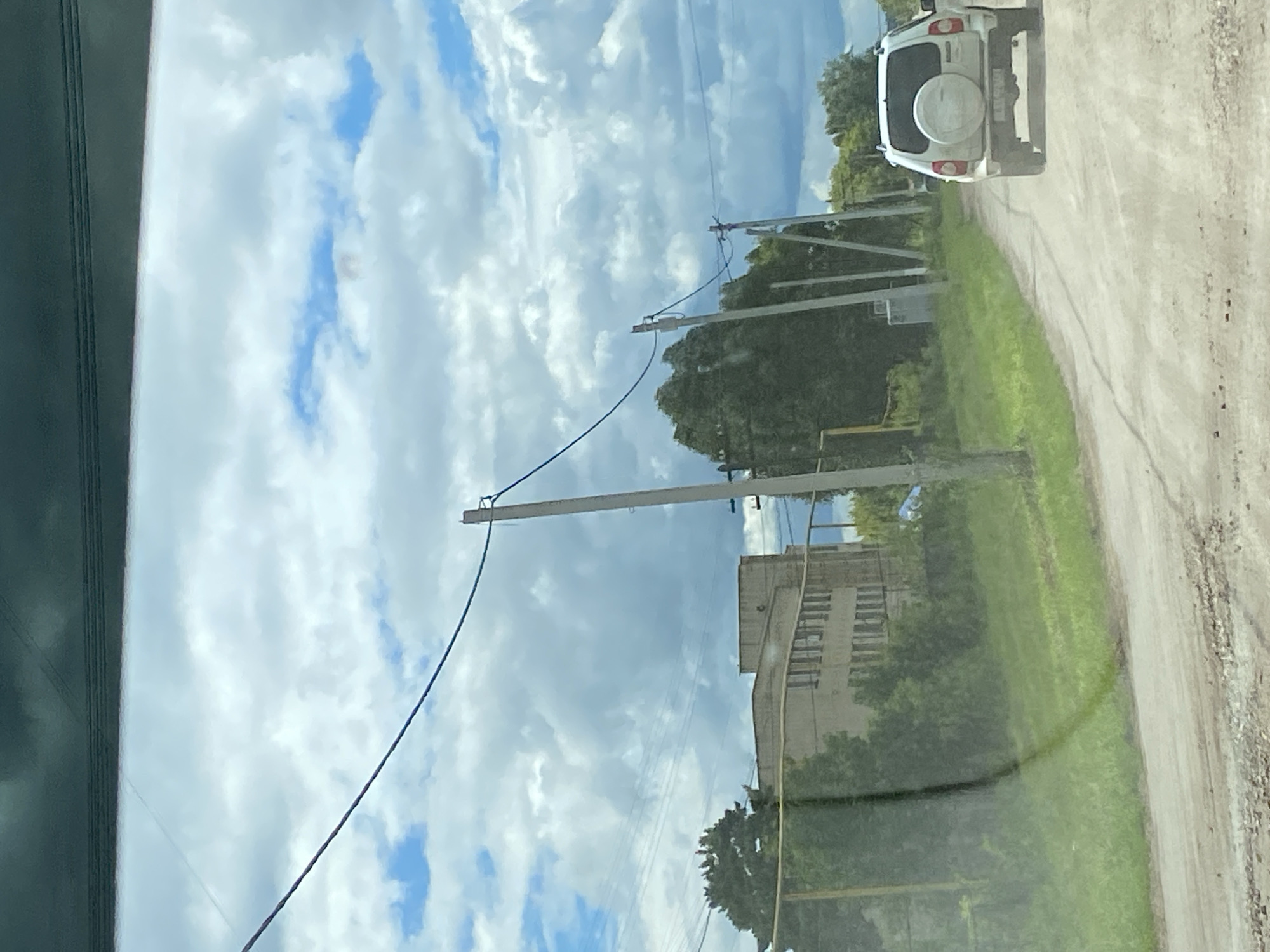 The image shows a white SUV driving on a dirt road in a rural area. The SUV is in the foreground, and the road is dusty. On the left side of the road, there is a building with a flat roof and several windows. There are also trees and power lines in the background. The sky is cloudy with patches of blue. The location appears to be outside of the city of Nadezhdino, Russia.