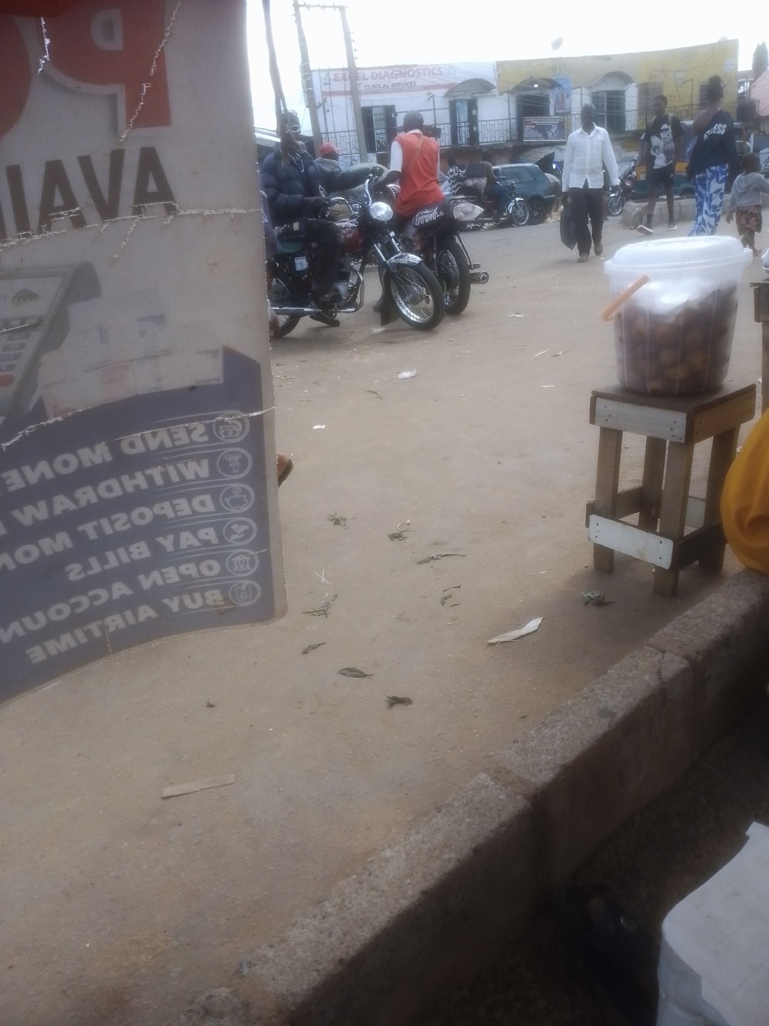 This image depicts a bustling street scene in Gwong, Nigeria, likely during daytime given the ambient light. The foreground is dominated by a large, torn poster with text partially visible. The text appears to be a service advertisement, listing "SEND MONEY," "WITHDRAW MONEY," "DEPOSIT MONEY," "PAY BILLS," "OPEN ACCOUNT," and "BUY AIRTIME." The poster is angled, showing some of its reflective surface.
In the mid-ground, several people are present, some on motorcycles. One person on a motorcycle wears a red vest and a helmet, appearing to be waiting or moving slowly. Another motorcycle rider is visible further back. A man in a white shirt walks with a dark bag, and a woman in a patterned blue and white outfit walks past him, followed by a young child. Another person in a dark shirt and pants is also visible.
The setting suggests a commercial or public area with shops in the background. One building has a sign that reads "EXCEL DIAGNOSTICS" and "CLINICAL SERVICES." Various motorcycles are parked or in motion, indicating transportation is a primary mode of movement. A large plastic tub filled with what appear to be small, round, fried food items sits on a rustic wooden stool in the right foreground, suggesting a food vendor or a snack stall. The ground is dusty and uneven, with some debris scattered around. The overall atmosphere appears active and typical of a lively market or transport hub.