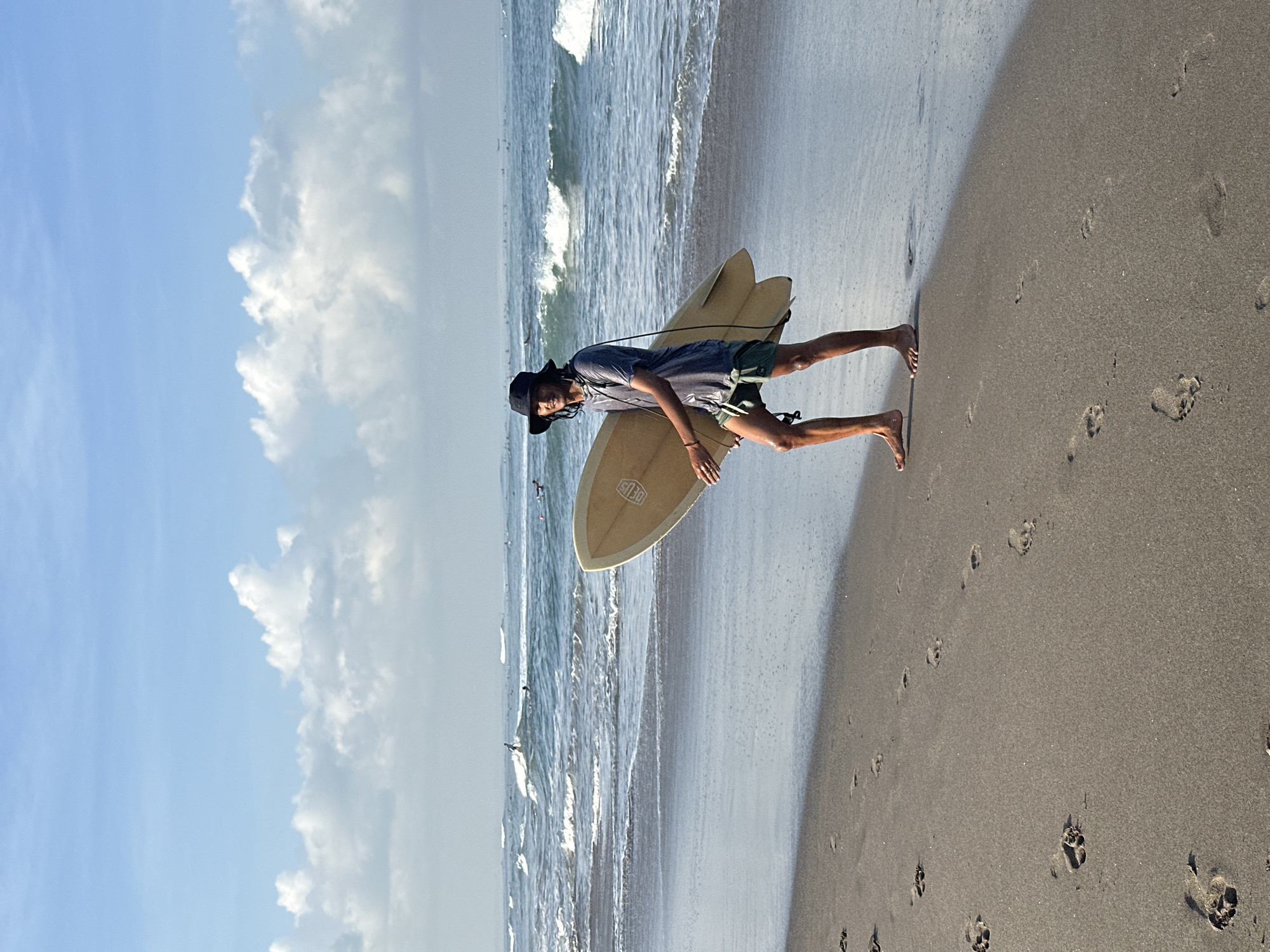 In the city of North Kuta, Indonesia, a young man is walking on a wet sandy beach carrying a light-colored surfboard under his right arm. He is wearing a dark bucket hat, a dark t-shirt, and green shorts. His left leg is slightly forward of his right, and his gaze is directed towards the ocean. The surfboard has a darker line around its perimeter and some markings on it. The ocean in the background has gentle waves, and several other surfers are visible in the distance, some riding waves. The sky is blue with scattered white clouds. Footprints are visible in the wet sand on the beach.