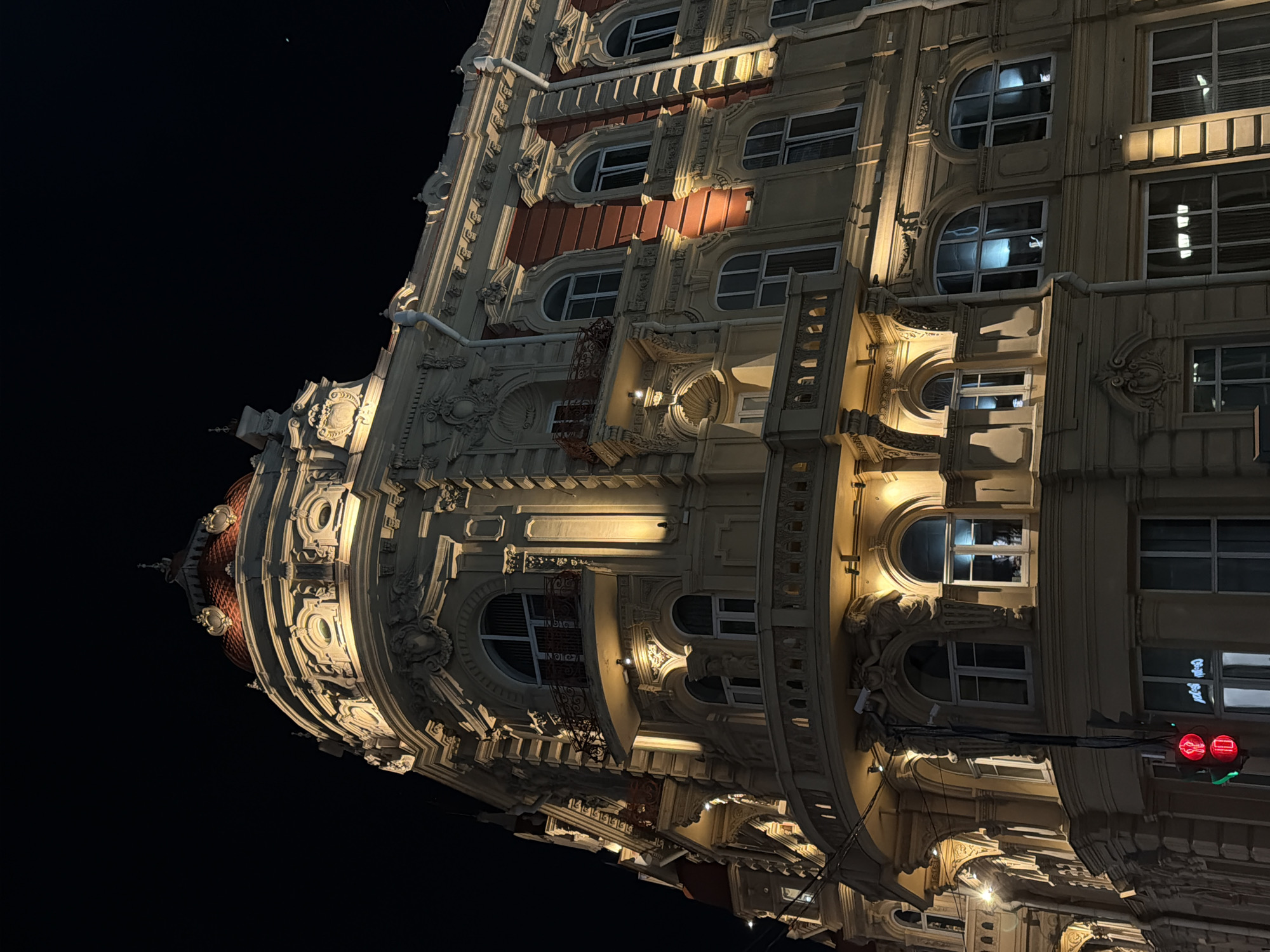 Here is a description of the image:
The image shows a building at night in Rostov-on-Don, Russia. The building is ornately decorated with sculptures and has a domed roof. The building is illuminated, and the sky is dark. There is a traffic light visible in the lower right corner of the image, showing a red light.