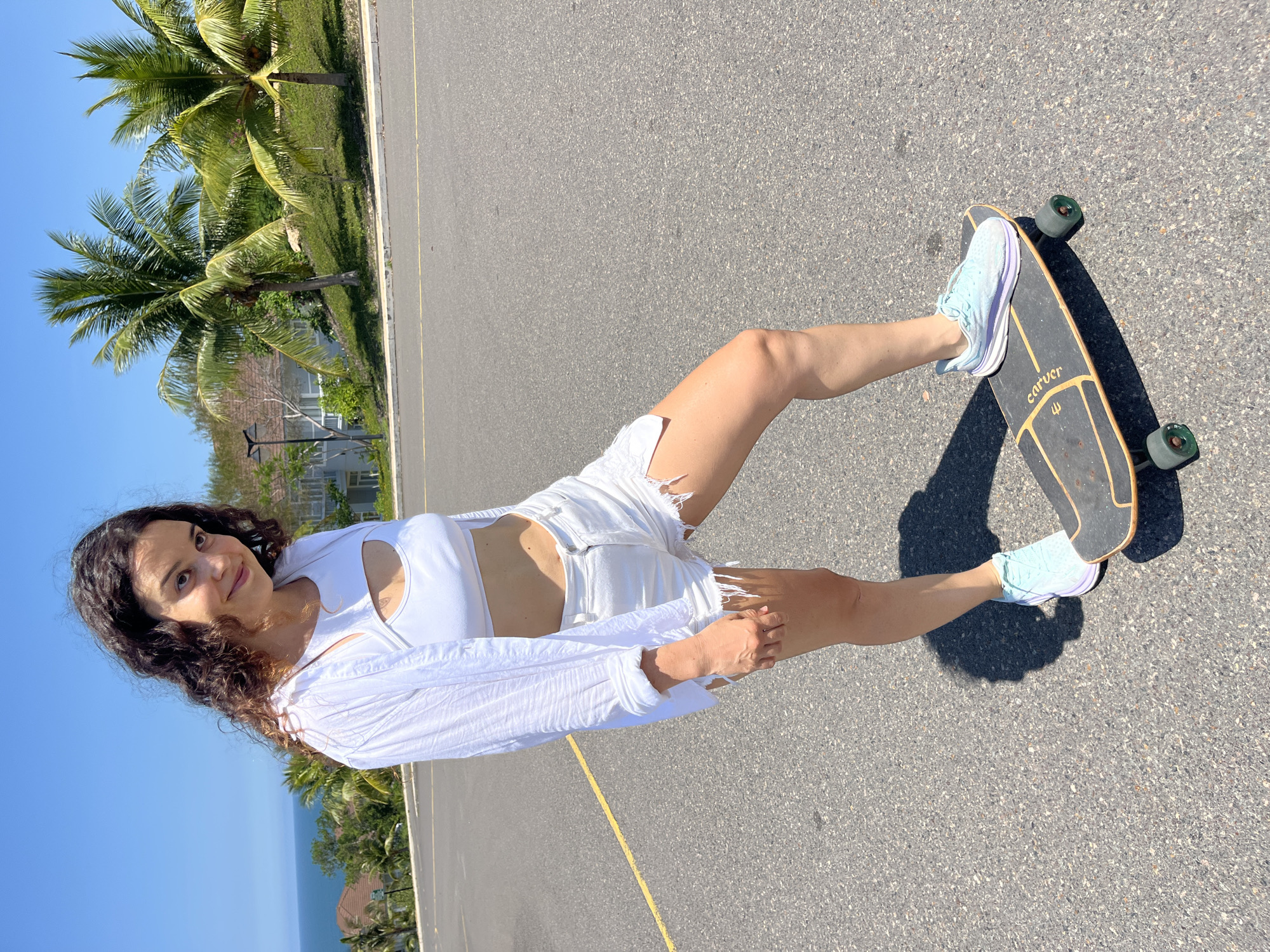 A young woman with long, curly brown hair stands on a skateboard on an asphalt surface, likely a parking lot, with a yellow line marking a lane. She is wearing a white crop top with a cut-out detail, distressed white denim shorts, and light blue and purple sneakers. A loose white long-sleeved shirt is draped over her shoulders. In the background, palm trees and buildings are visible, with the ocean in the distance. The scene is brightly lit by sunlight, suggesting a clear day. The location is identified as Phan Thiết, Vietnam.