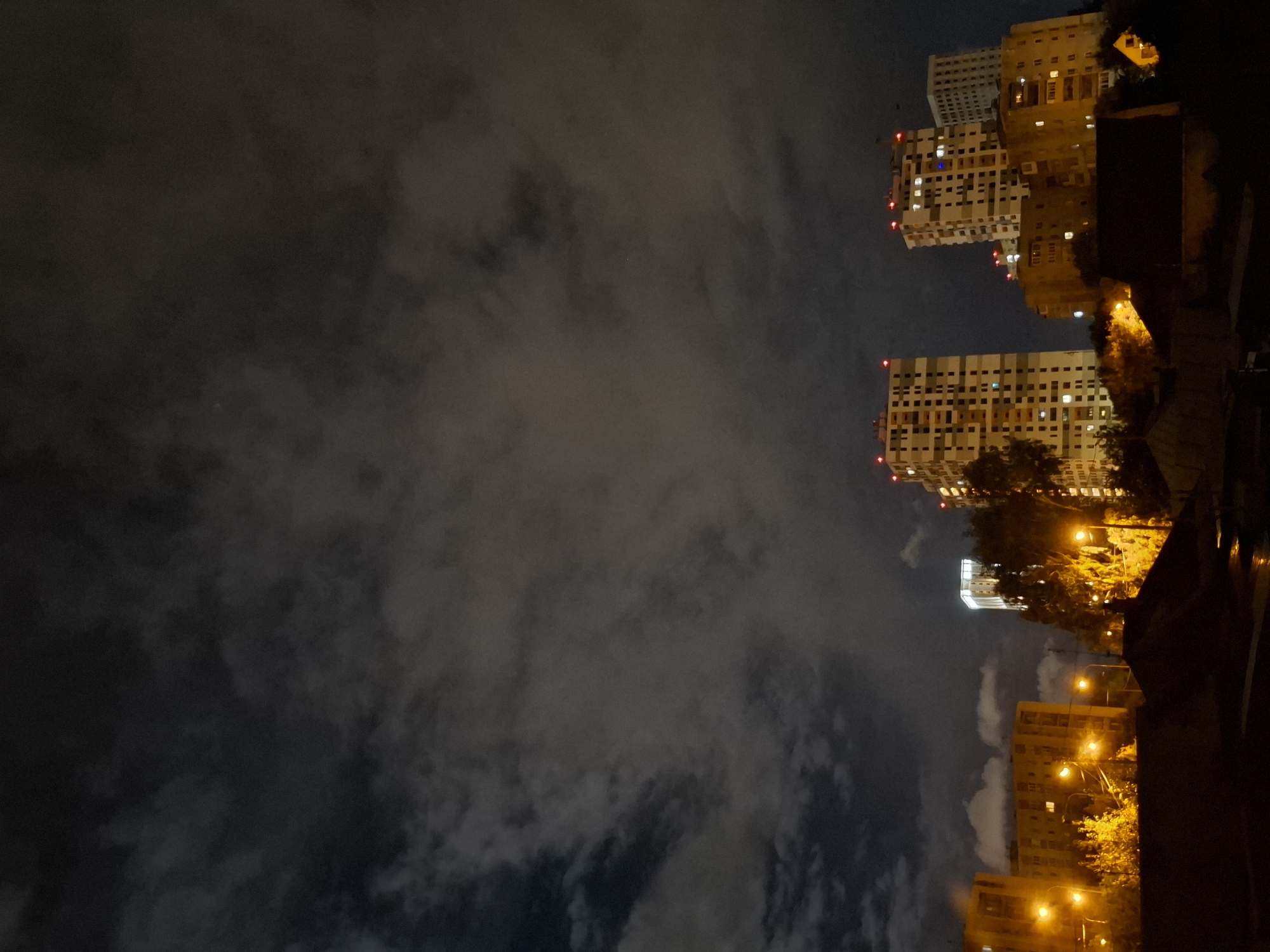 This is a low-angle, nighttime photograph of a cityscape, likely in Tyumen, Russia, given the context. The sky is filled with dramatic, dark clouds, with some lighter patches suggesting moonlight filtering through. In the mid-ground and background, several multi-story buildings are visible.
The most prominent structures are tall, modern apartment buildings, characterized by their numerous windows, many of which are illuminated with warm, yellow light. Some of these buildings have red lights at their very tops, possibly for aviation safety. One building in the center-left is significantly shorter and appears to be an older residential block, with yellow-toned walls and illuminated windows.
In the foreground and mid-ground, streetlights cast a strong, warm glow on the surrounding trees and vegetation, creating a vibrant, almost autumnal effect despite the nighttime setting. The lower portion of the image shows darker, silhouetted structures, possibly rooftops or lower buildings, with some indistinct shapes suggesting foliage or debris. The overall impression is of a residential urban area at night, with the city lights contrasting against the dark, cloudy sky. There are no visible people or vehicles in the frame.