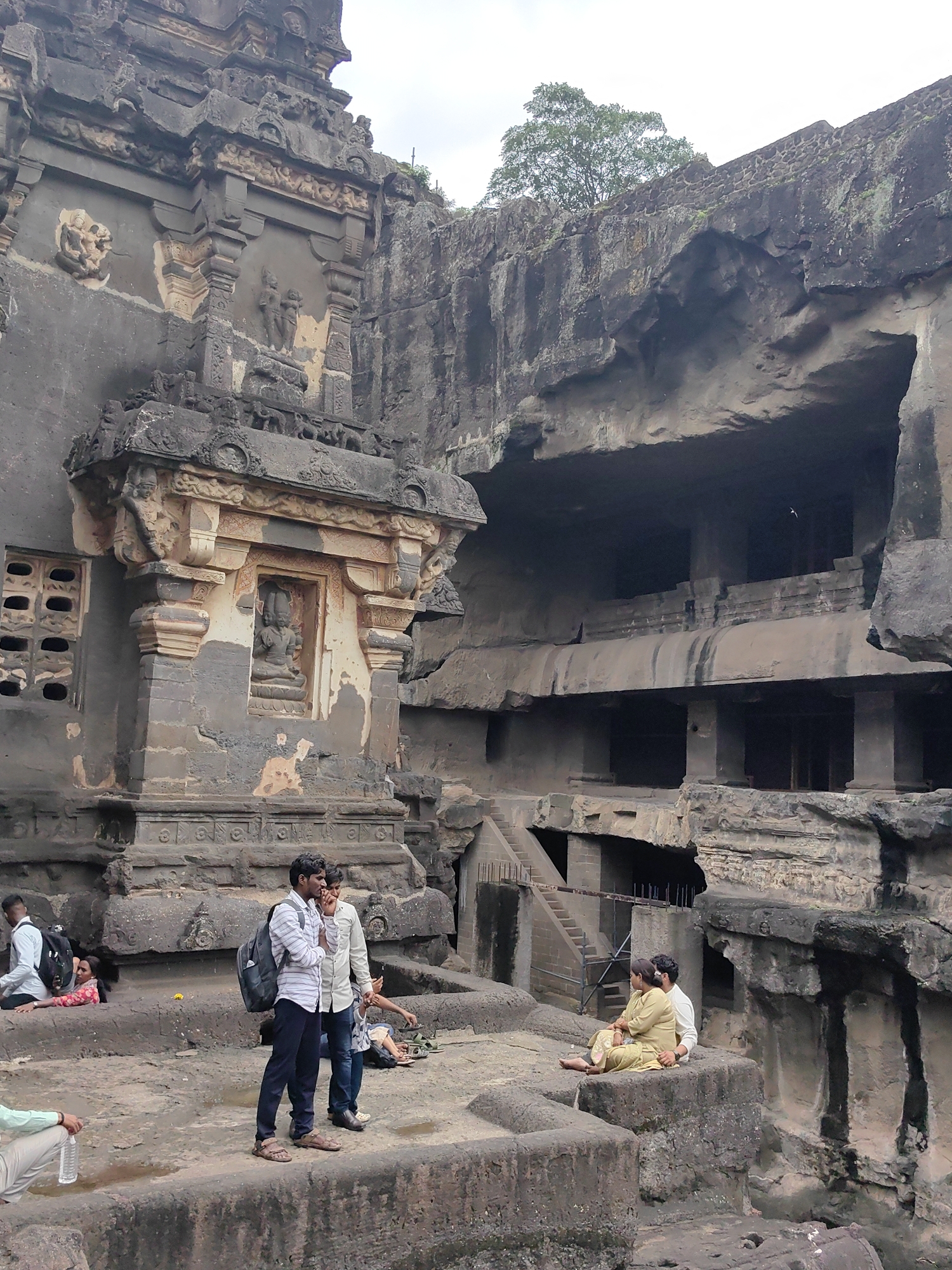 The image shows the Ellora Caves in Ellora, India, a UNESCO World Heritage site known for its rock-cut architecture. The scene depicts visitors interacting within the ancient cave complex. In the foreground, several people are present. Two men stand side-by-side, one wearing a striped shirt and dark pants, the other a light-colored shirt and blue jeans. Both have backpacks. To their left, a woman in a pink and white patterned top is lying down. Further to the left, a man in a light green shirt and light-colored pants is seated, holding a plastic water bottle. In the middle ground, two people are seated on a rock ledge. The person on the left is wearing a yellow garment, and the person on the right, partially obscured, appears to be wearing a white shirt. Behind them, a massive rock-cut structure, characteristic of the Ellora Caves, dominates the background. The structure features intricate carvings, architectural elements, and openings into deeper parts of the cave. A tree is visible growing from the top of the rock formation. The overall environment is rocky and ancient, with the weathered stone and the presence of both historical architecture and modern-day visitors.