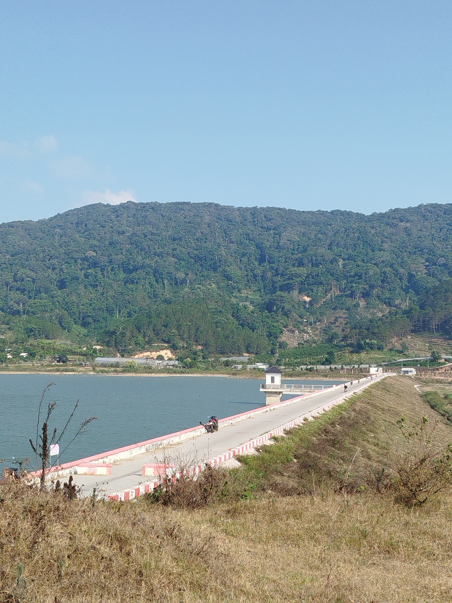 The image shows a reservoir in Đông Hồ, Vietnam. A paved road extends across the water, bordered by a red and white painted curb. A small, two-story tower stands on the reservoir side of the road. A mountain covered in dense forest forms the background. A motorcycle is parked on the road, and other people are present on the road further away. The foreground is dominated by dry grass and scrub vegetation. The sky is clear and blue.