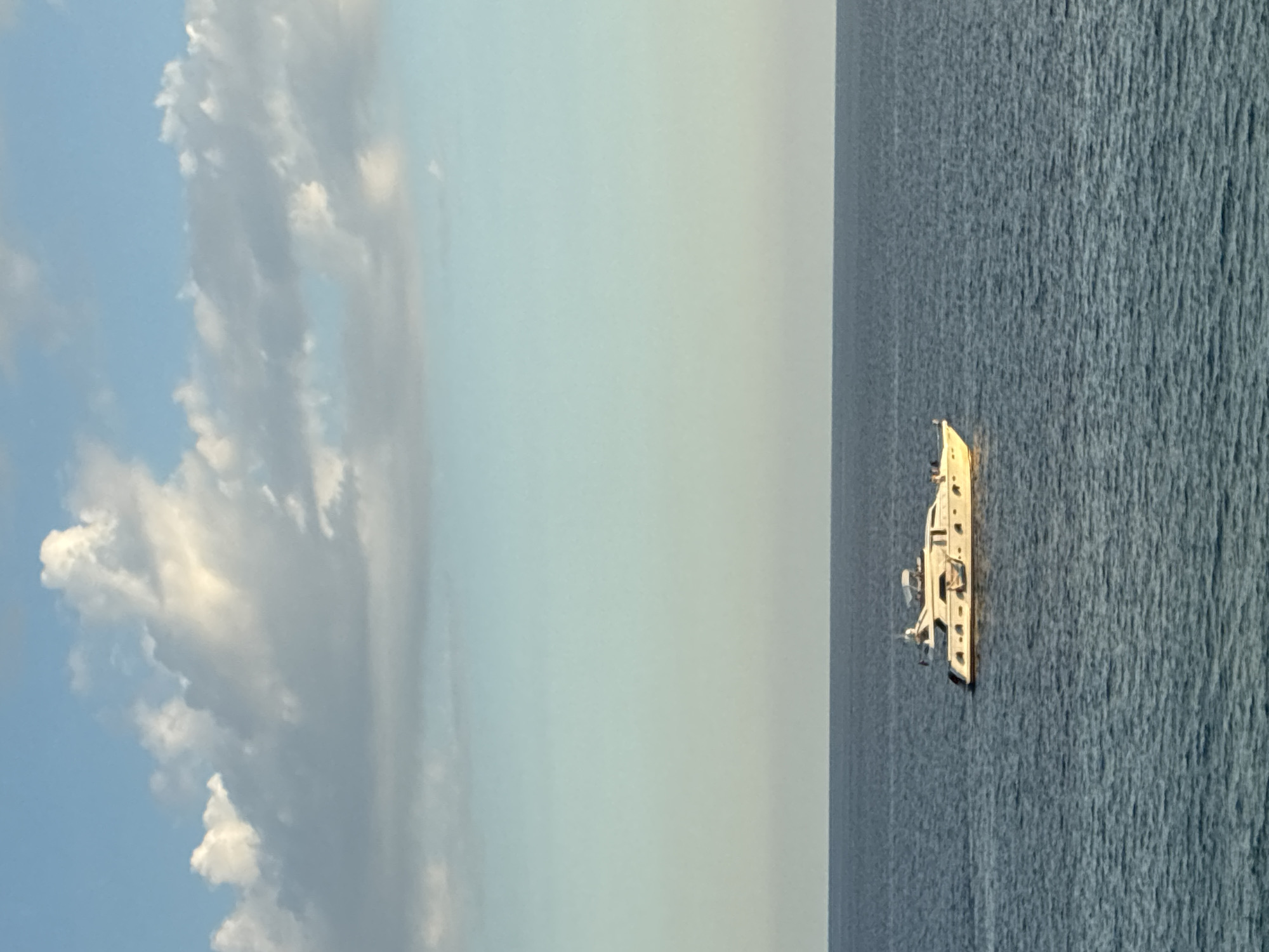 The image depicts a white yacht sailing on calm, dark blue waters. The yacht has multiple levels and appears to be a luxury vessel. In the background, the sky is a light blue with scattered white clouds, some of which are illuminated by sunlight. The horizon line is clearly visible, separating the sea from the sky. There are no buildings or landforms visible in the image, suggesting an open water setting. The image is a photograph taken from a distance, likely from another boat or a coastal vantage point. The location is not specified in the image itself, and therefore cannot be identified as Alanya, Turkey.