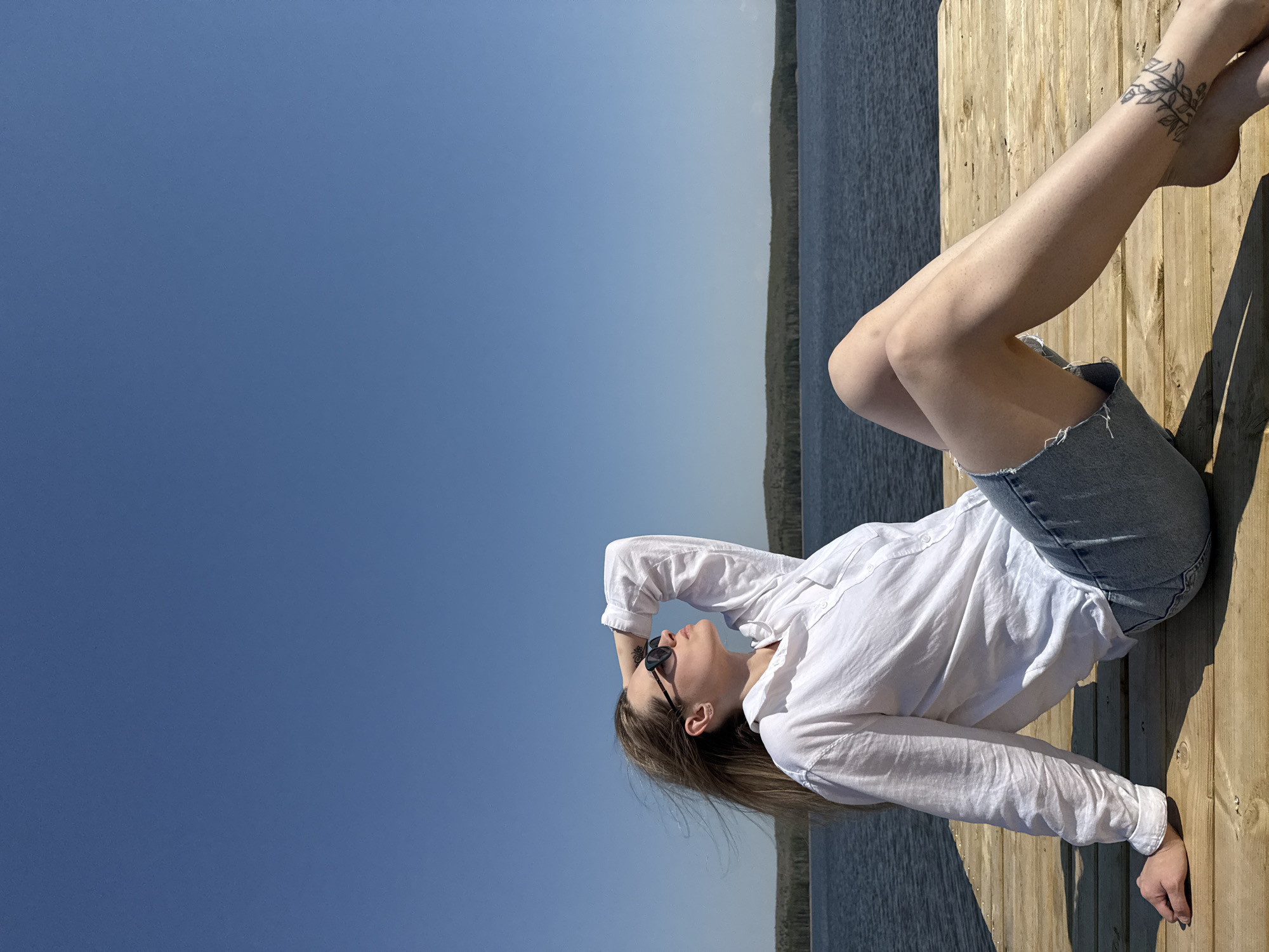 A young woman is lying on her side on a wooden dock, with a body of water and horizon visible in the background. The woman is wearing a white, long-sleeved shirt and denim shorts. She has sunglasses on and her hair is light brown. Her right hand is resting on her head and her left hand is near her waist. The woman has a visible tattoo on her left ankle. The sky is clear and blue. The scene appears to be in the city of Chaikovsky, Russia based on information that you provided.