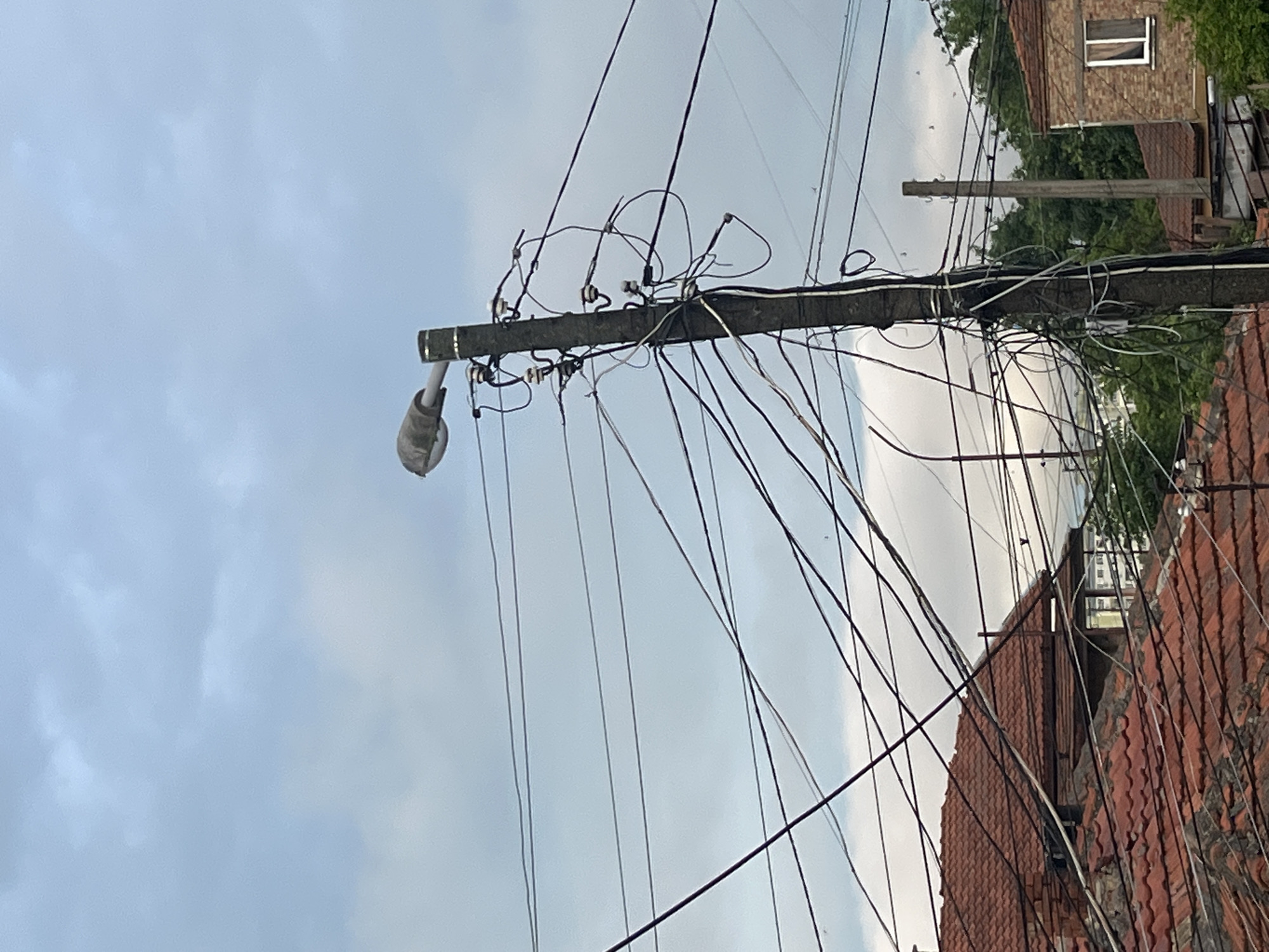 The image, rotated 90 degrees clockwise, captures a densely wired utility pole against an overcast sky. The sturdy, dark-colored pole stands prominently, featuring a grey street light fixture extending from its left side. A complex network of numerous black and grey electrical and communication wires emanates from the pole, crossing the frame in various directions. Many wires appear loosely arranged, tangled, or looped around attachments, where several insulators are visible.
In the background, to the right of the main pole, sections of residential buildings with distinctive red-tiled roofs are visible. Above these roofs, a brown brick building with a white-framed, double-paned window can be seen, partially obscured by green trees and foliage. A second, thinner utility pole is also present further in the background. The sky is a light grey with diffuse clouds, indicating a daytime setting without direct sunlight. There is no visible text or human activity in the scene.