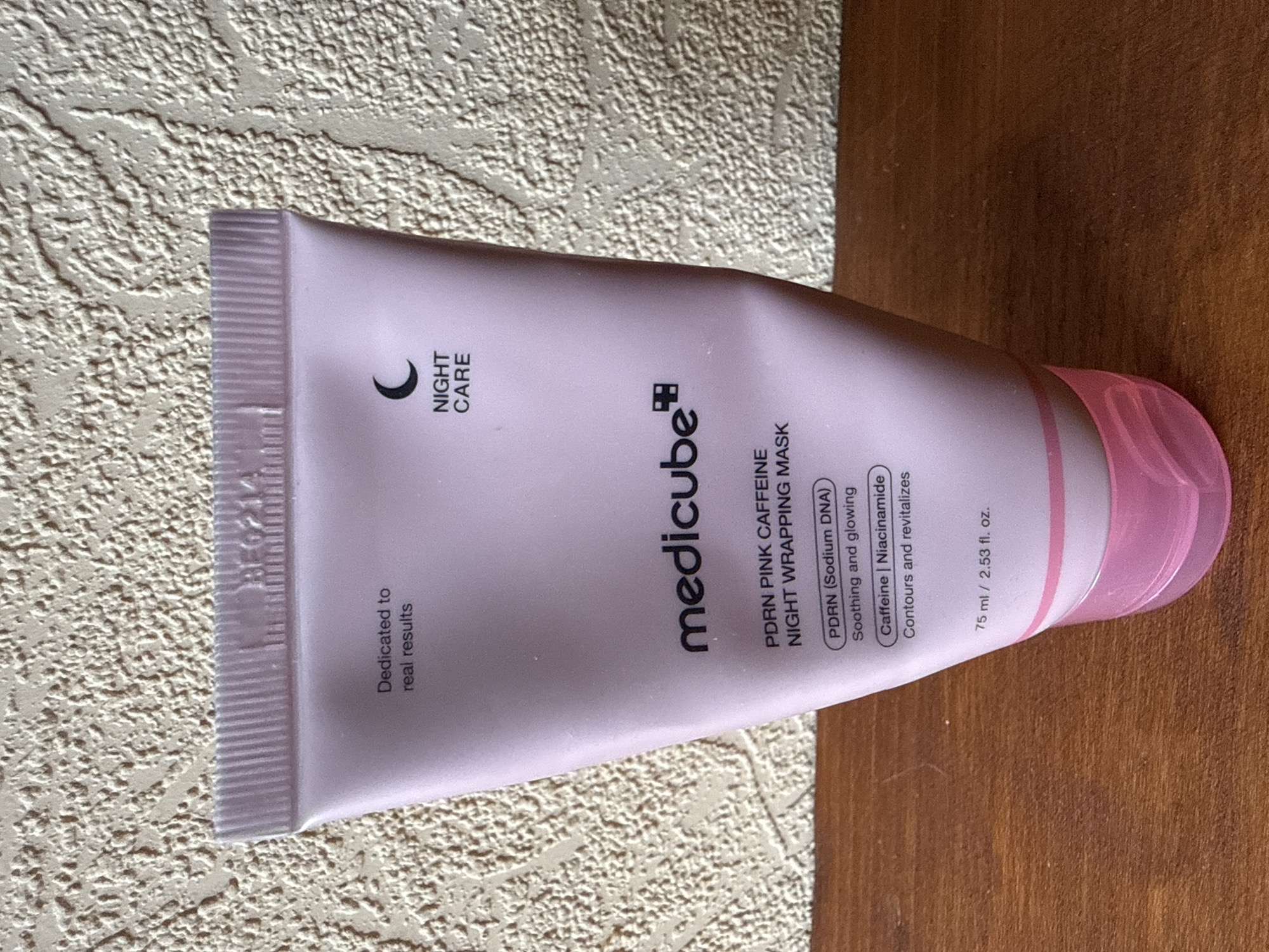 The image shows a pink tube of Medicube PDRN Pink Caffeine Night Wrapping Mask. The tube is standing upright on a wooden surface against a textured, light-colored background. Text on the tube reads "Dedicated to real results," "NIGHT CARE," "medicube," and "PDRN PINK CAFFEINE NIGHT WRAPPING MASK." Below this, further details include "PDRN (Sodium DNA) Soothing and glowing" and "Caffeine | Niacinamide Contours and revitalizes." The volume is listed as "75 ml / 2.53 fl. oz." There are no people or actions visible in the image. The context of Okhtyrka, Ukraine, is not discernible from the image itself.